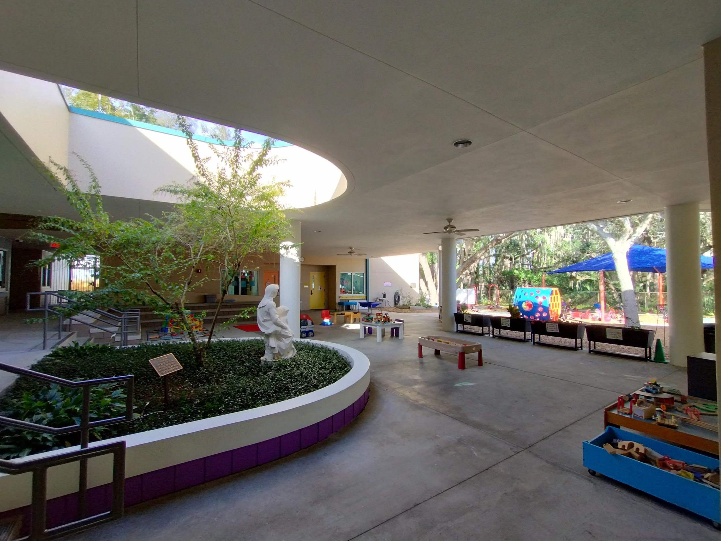 Indoor playground area with a statue of a person holding a child, surrounded by plants, colorful toys, and outdoor play structures visible through windows, with benches and shade structures.