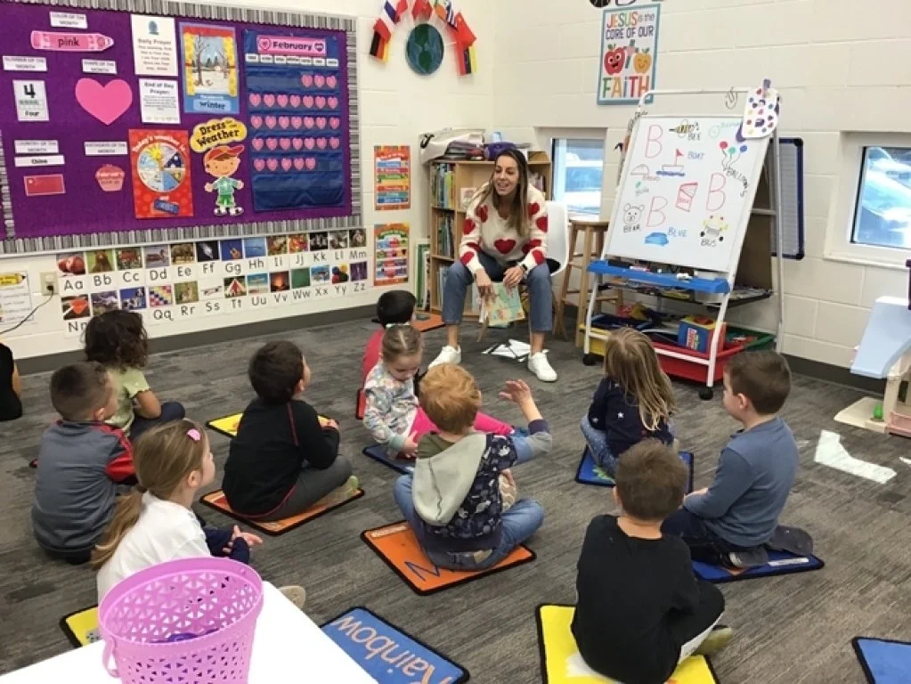 A woman sitting on a chair in a classroom with young children sitting on colorful mats on the floor in front of her. The classroom has educational posters, a whiteboard with drawings and letters, and colorful decorations including a rainbow and a heart-themed bulletin board.