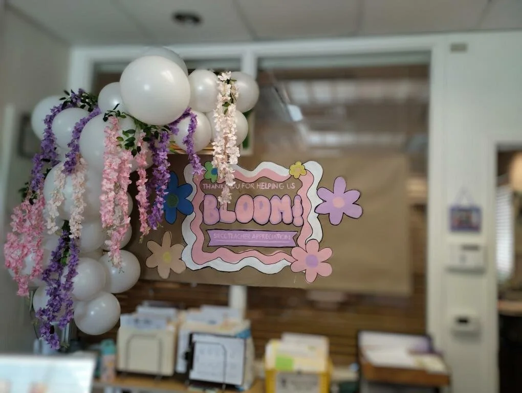 Decorated classroom display with a paper sign saying "BLOOM!" surrounded by white balloons and pink and purple floral decorations, with a background of an indoor room.