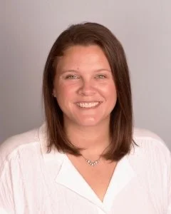 A woman with shoulder-length brown hair smiling, wearing a white top, against a neutral background.