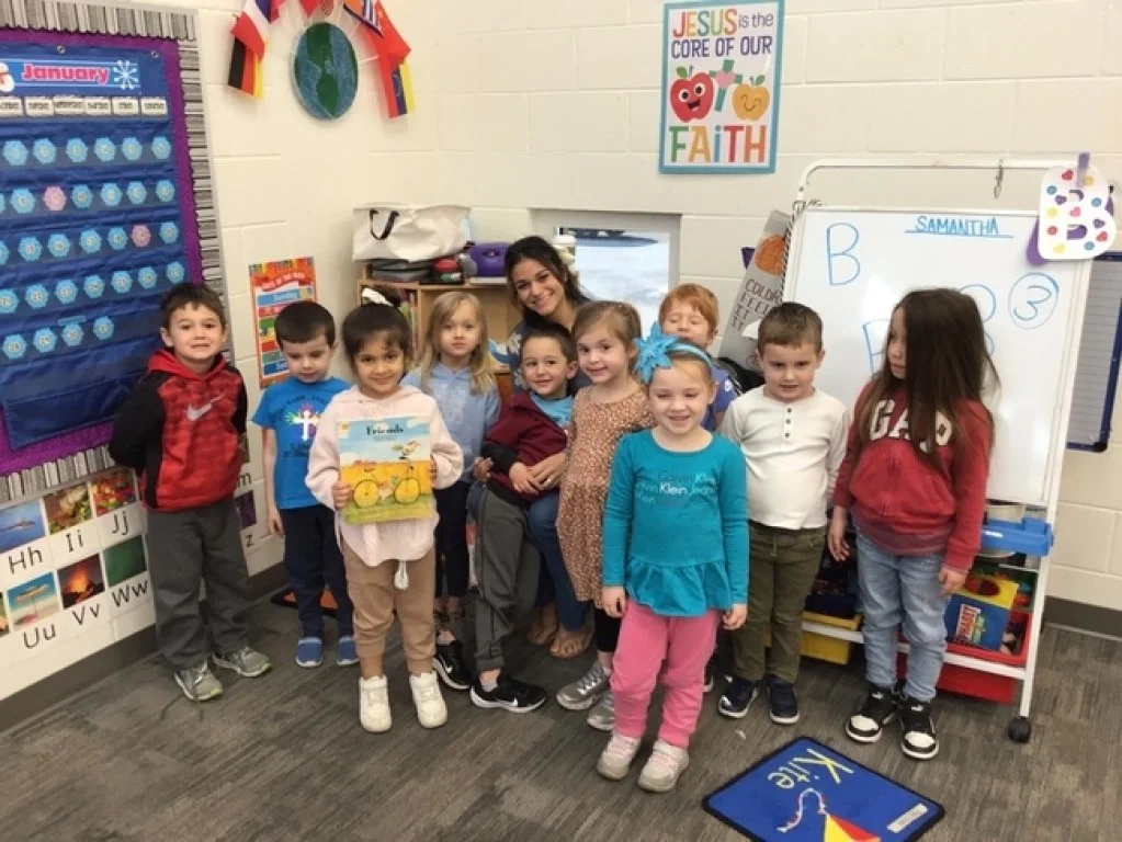 Group of young children and a teacher posing in a classroom with educational decorations and a whiteboard.