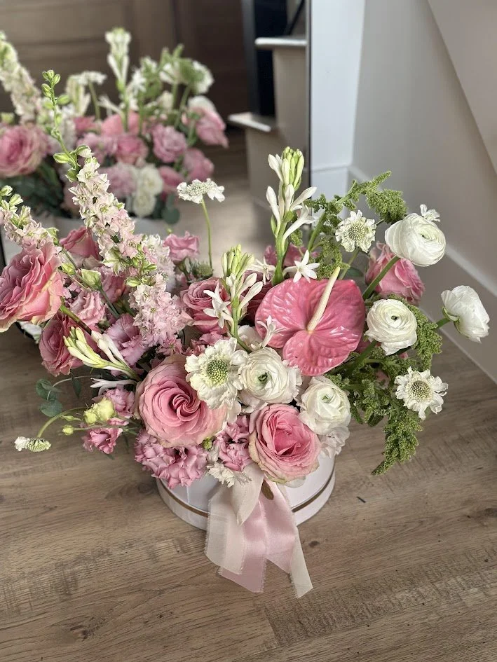 A pink and white floral arrangement with roses, anthuriums, and lilies in a round white vase with a pink ribbon on a wooden table.