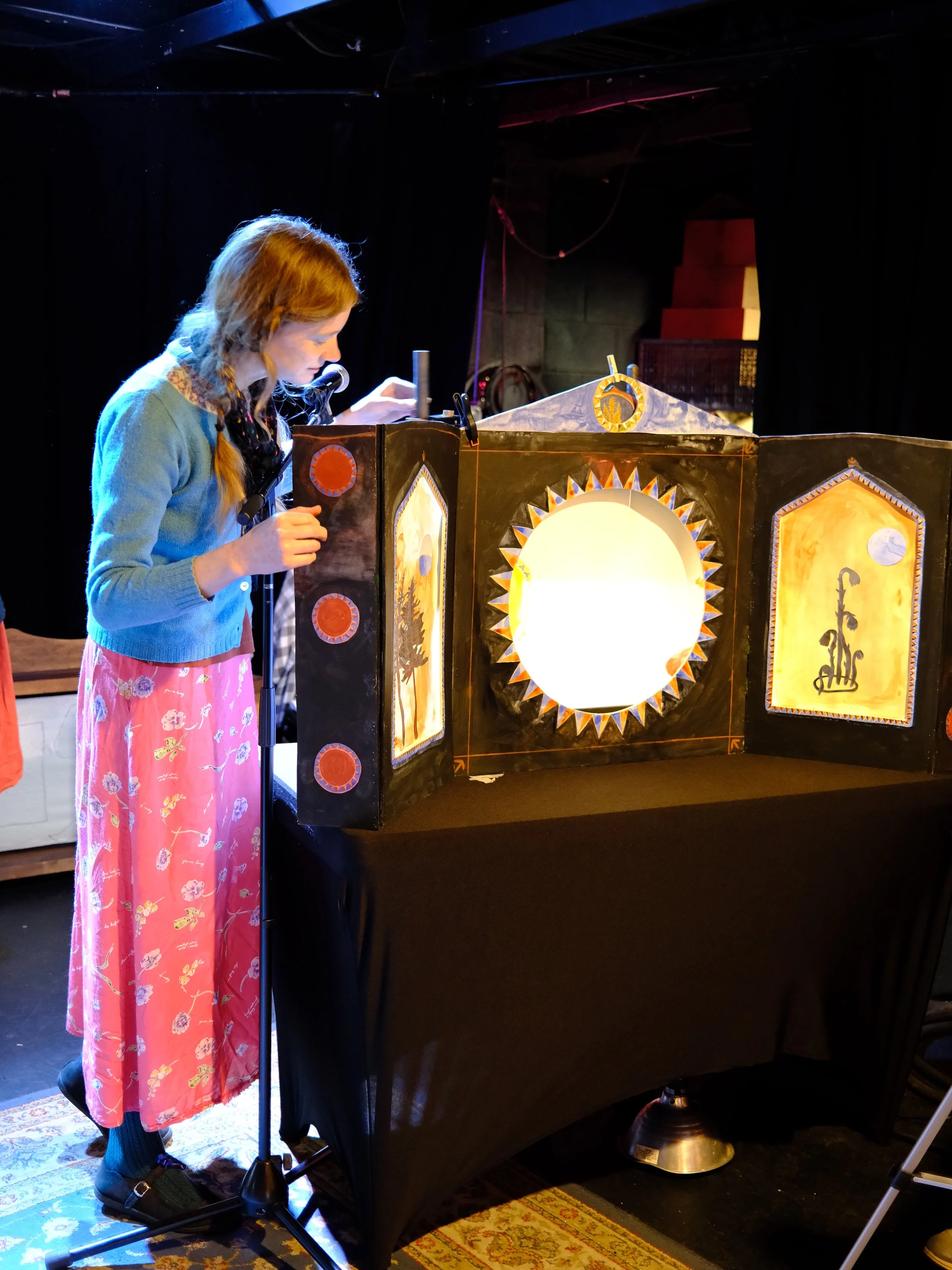 A young girl with red hair standing beside a puppetry stage, focusing on a puppet performance with a lighted backdrop, in a dark theater setting.