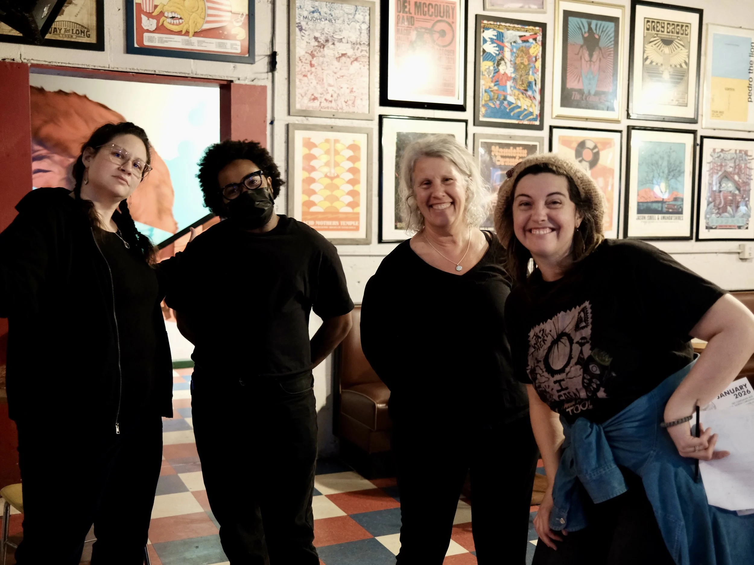 Four women standing together indoors in front of a wall with framed posters. Two women on the left are wearing dark clothing, one with glasses and a mask. The two women on the right are smiling, one with a gray hair and the other with a curly hair and a hat.