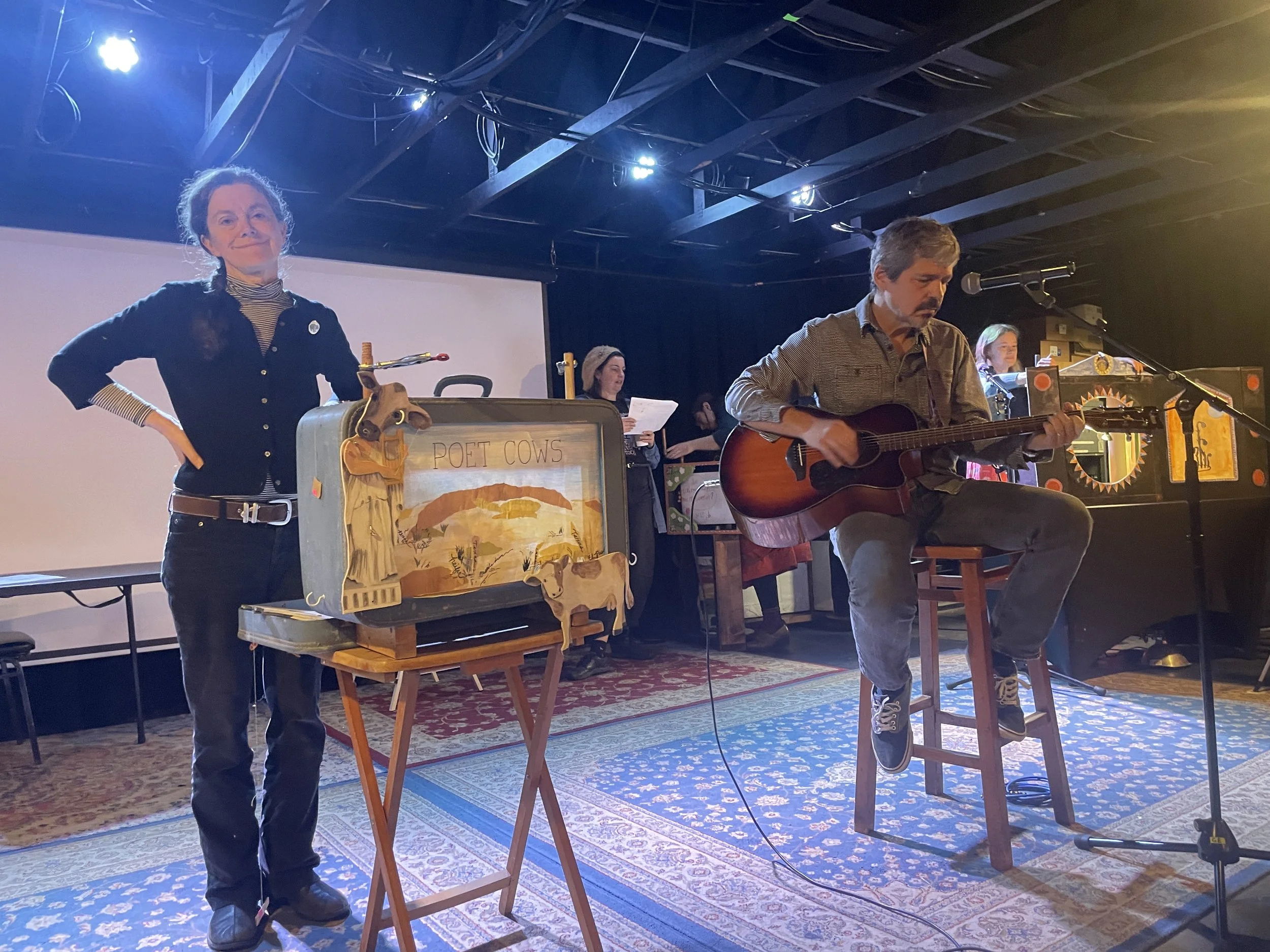 A man is playing an acoustic guitar while sitting on a stool, with two women and a puppet show setup including a puppet in front labeled 'Poet Cows'. The scene appears to be a performance or storytelling event in a venue with stage lighting and a backdrop.