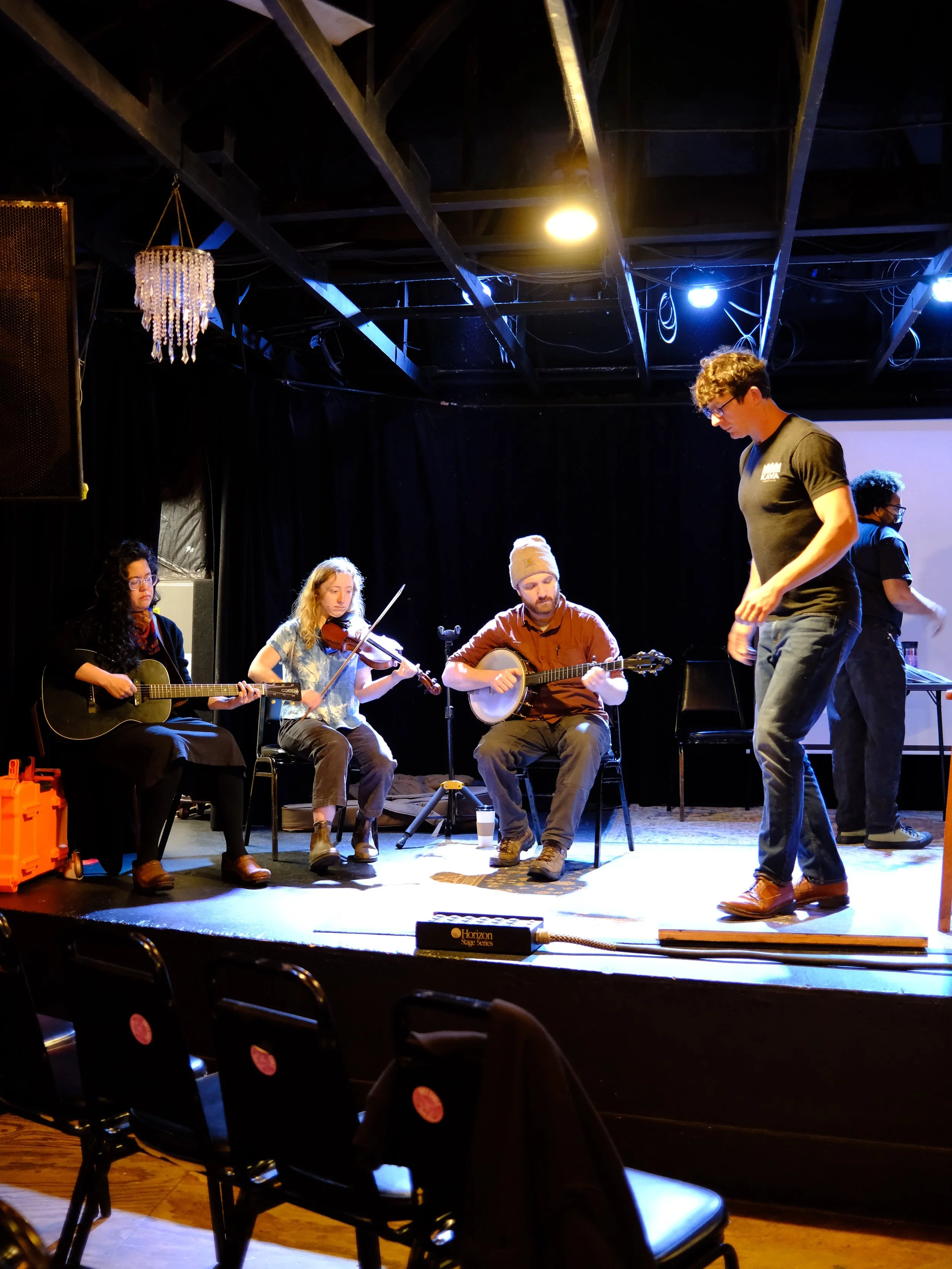 Musicians practicing on a small stage, including a woman with an acoustic guitar, a woman with a violin, a man with a banjo, and a man walking on stage, in a dimly lit room with black curtains and acoustic lighting.