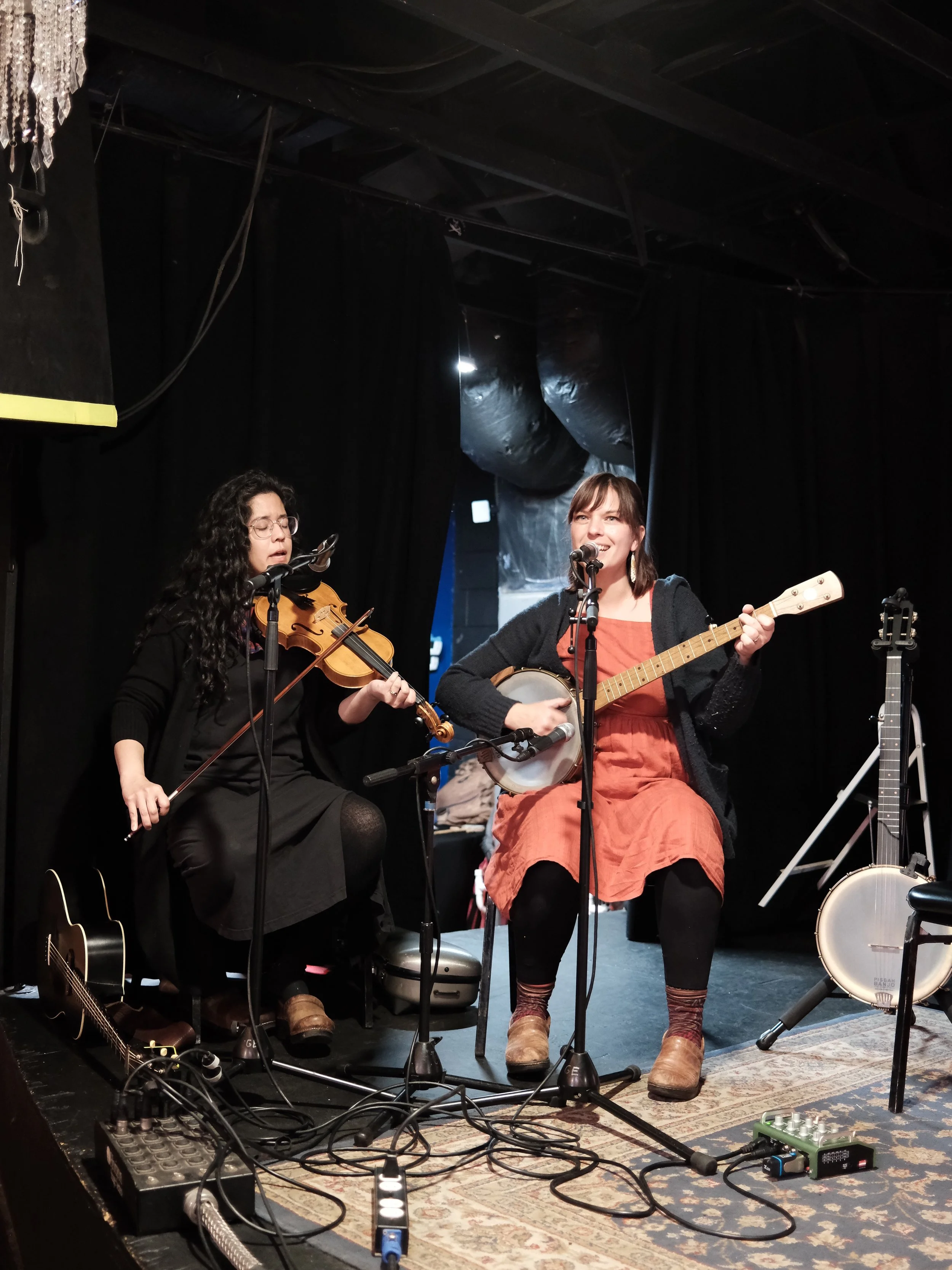 Two women performing on stage with musical instruments, a violin and a banjo, in a dark room with black curtains.
