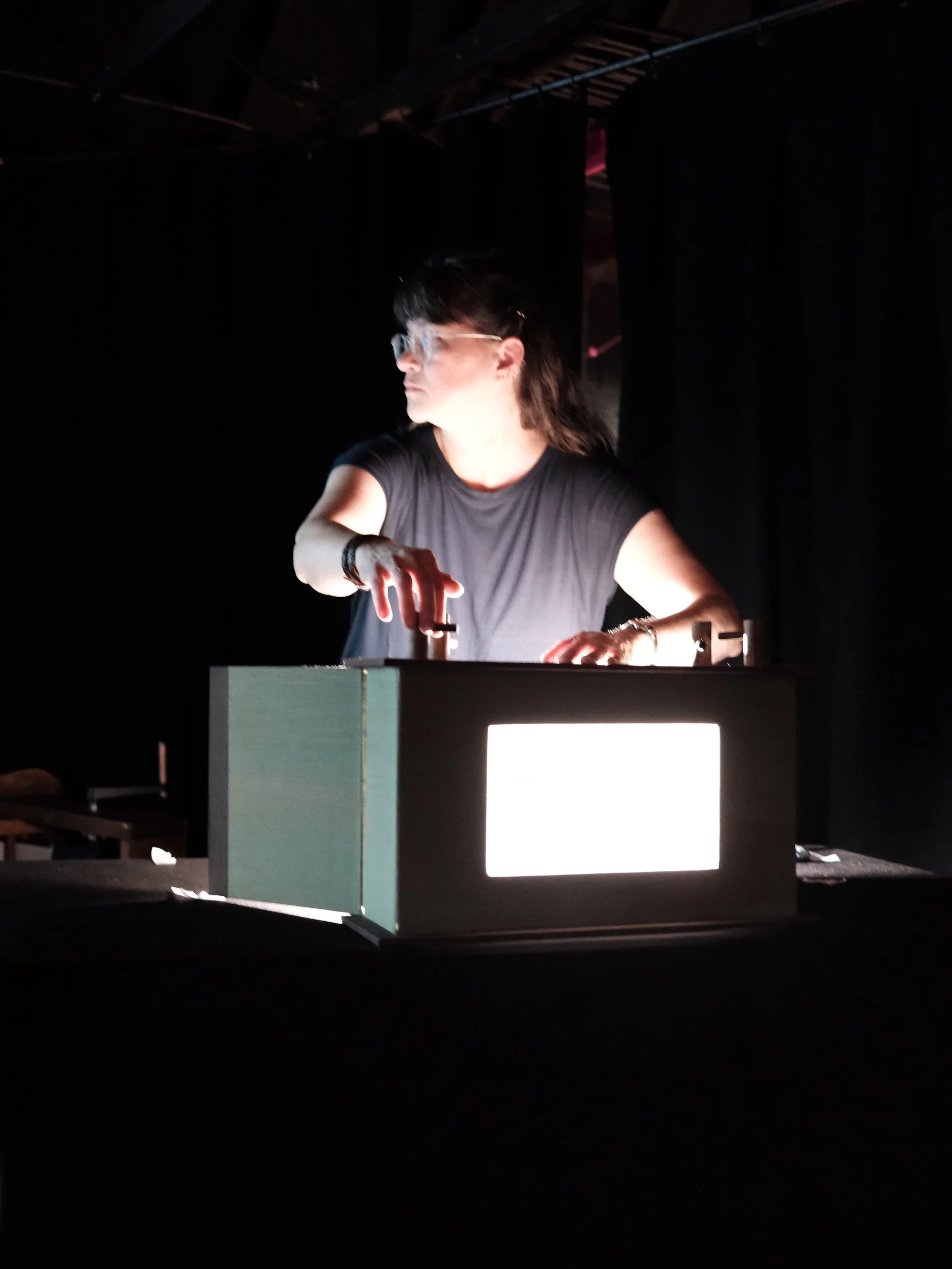 A woman with glasses working at a DJ console on a dark stage.