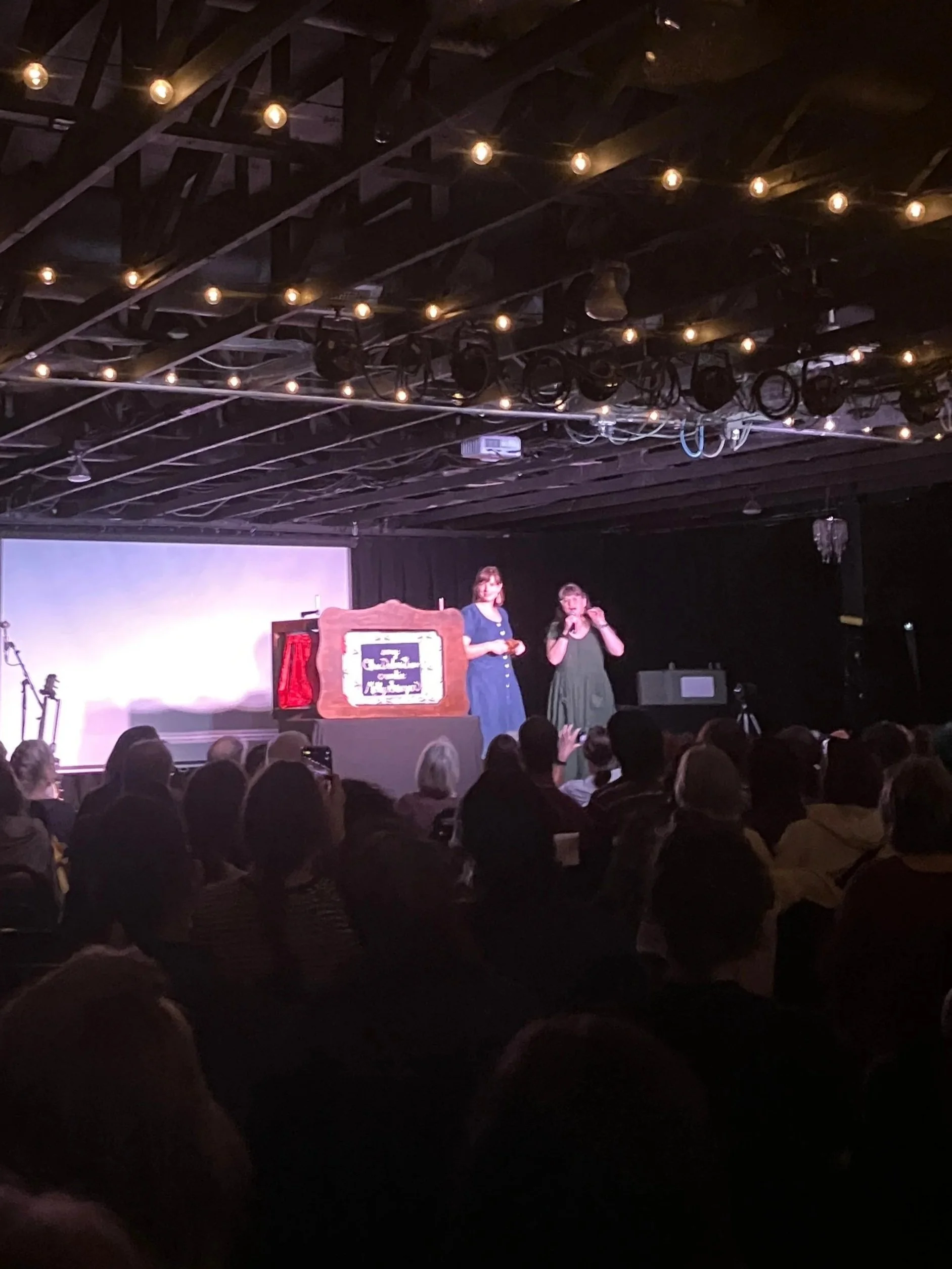 Audience watching a stage performance with two women standing in front of a puppet theater backdrop and a large screen behind them, all inside a dimly lit venue with string lights hanging from the ceiling.