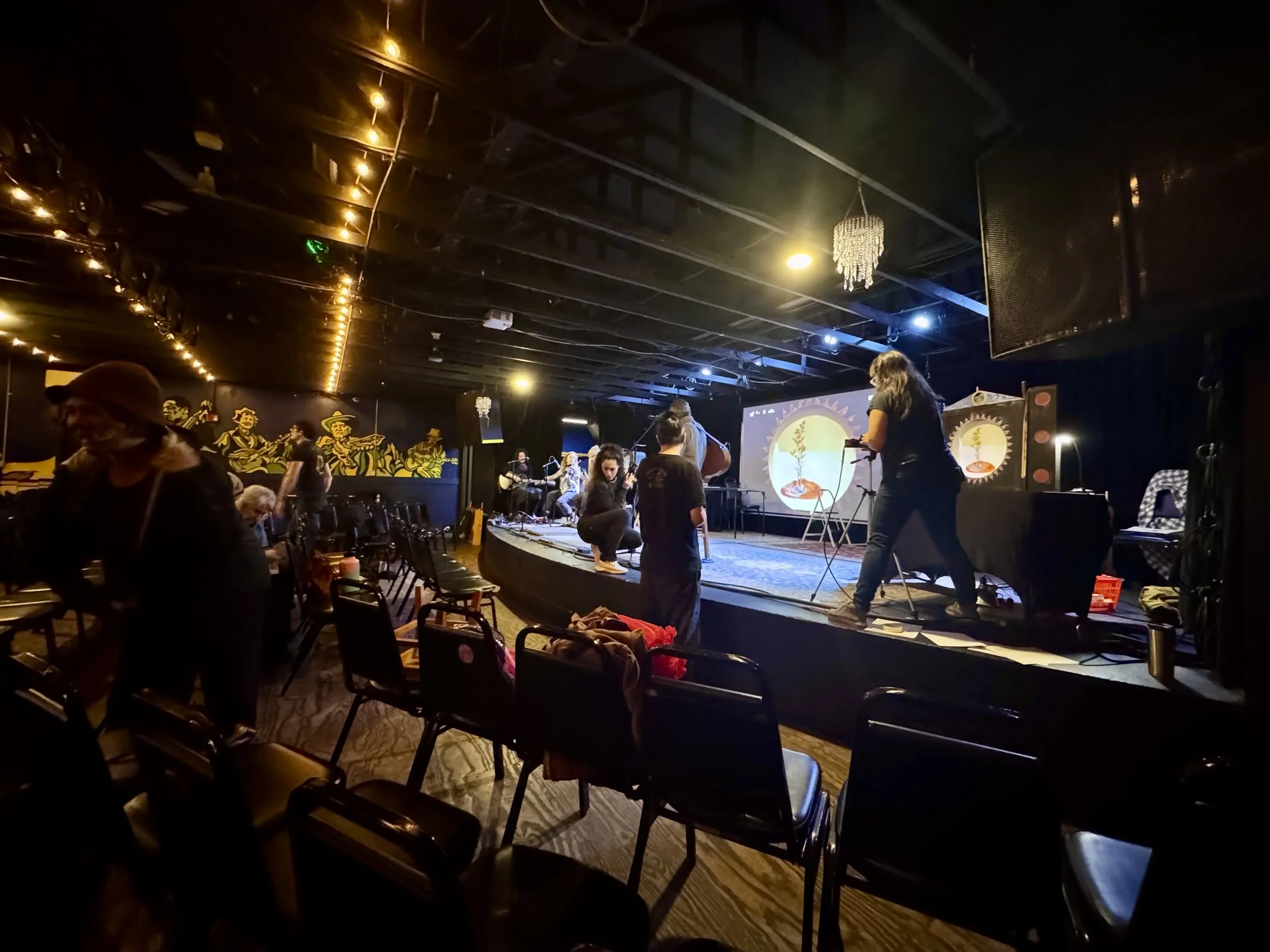Dimly lit performance venue with a small stage where musicians are setting up instruments. Behind them is a screen with a colorful, abstract graphic. The ceiling has warm string lights, and the background wall features illustrations of musicians.