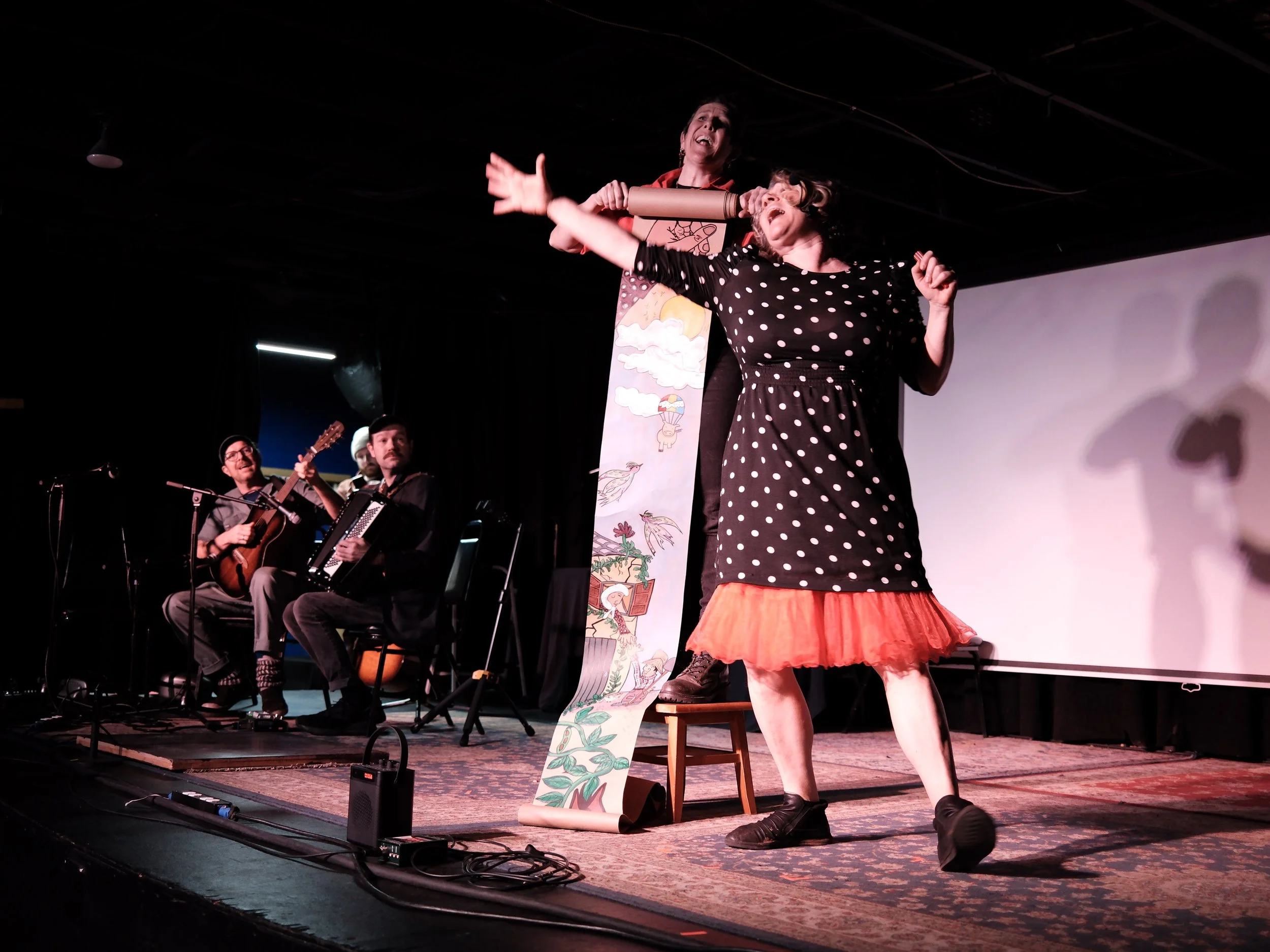 Two women are performing on stage, one is standing and the other is sitting on a stool, with musicians playing guitar and accordion in the background.