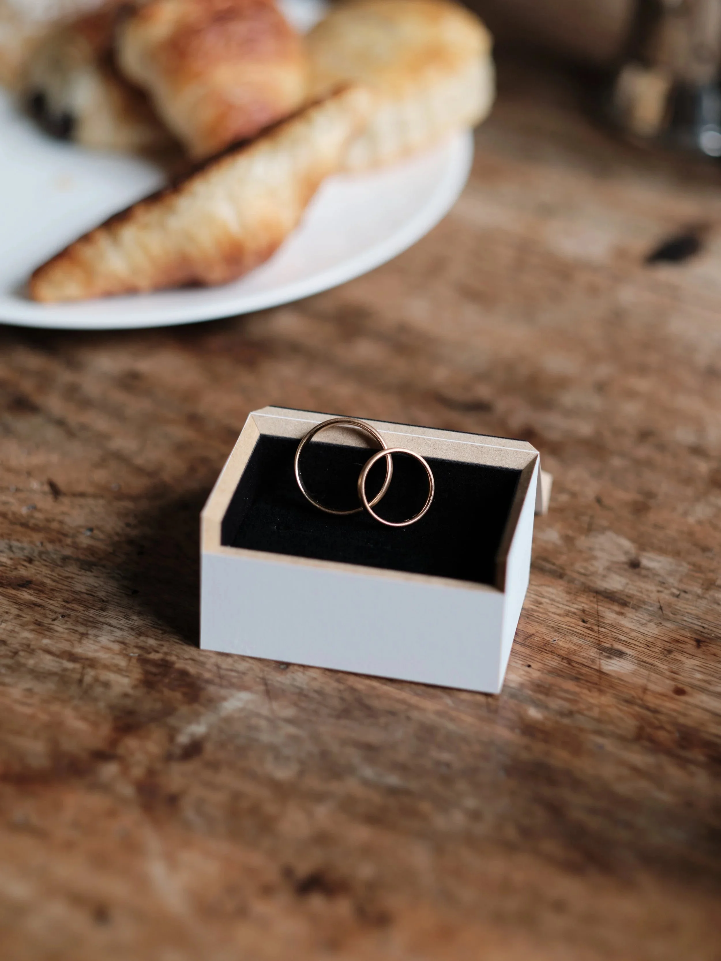 Zwei goldene Eheringe in einer schwarzen Samtschachtel auf einem Holztisch, im Hintergrund eine weiße Platte mit Gebäck.