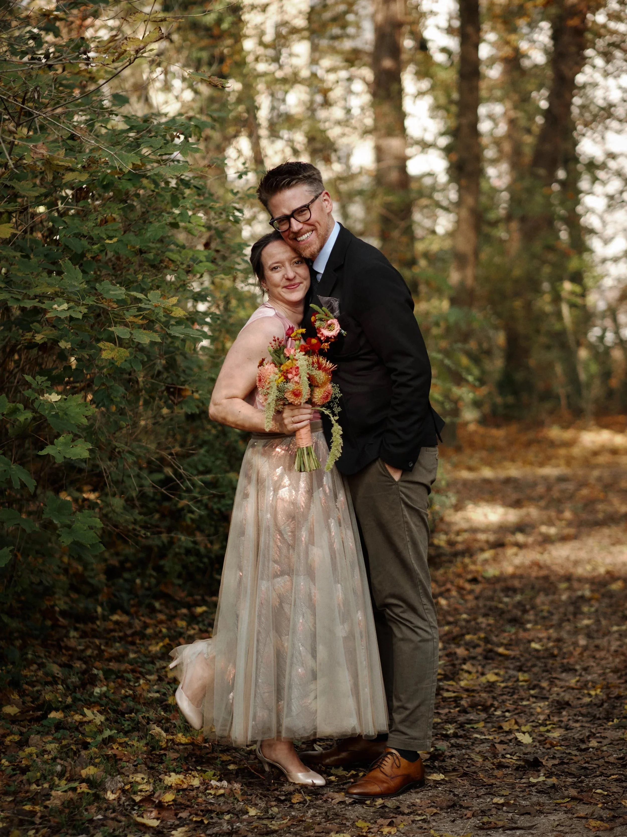 Hochzeitspaar umarmt sich. Außenportrait von Braut und Bräutigam. Die Frau trägt ein langes, zartes Kleid und hält einen Blumenstrauß, der Mann trägt eine schwarze Jacke und braune Schuhe.