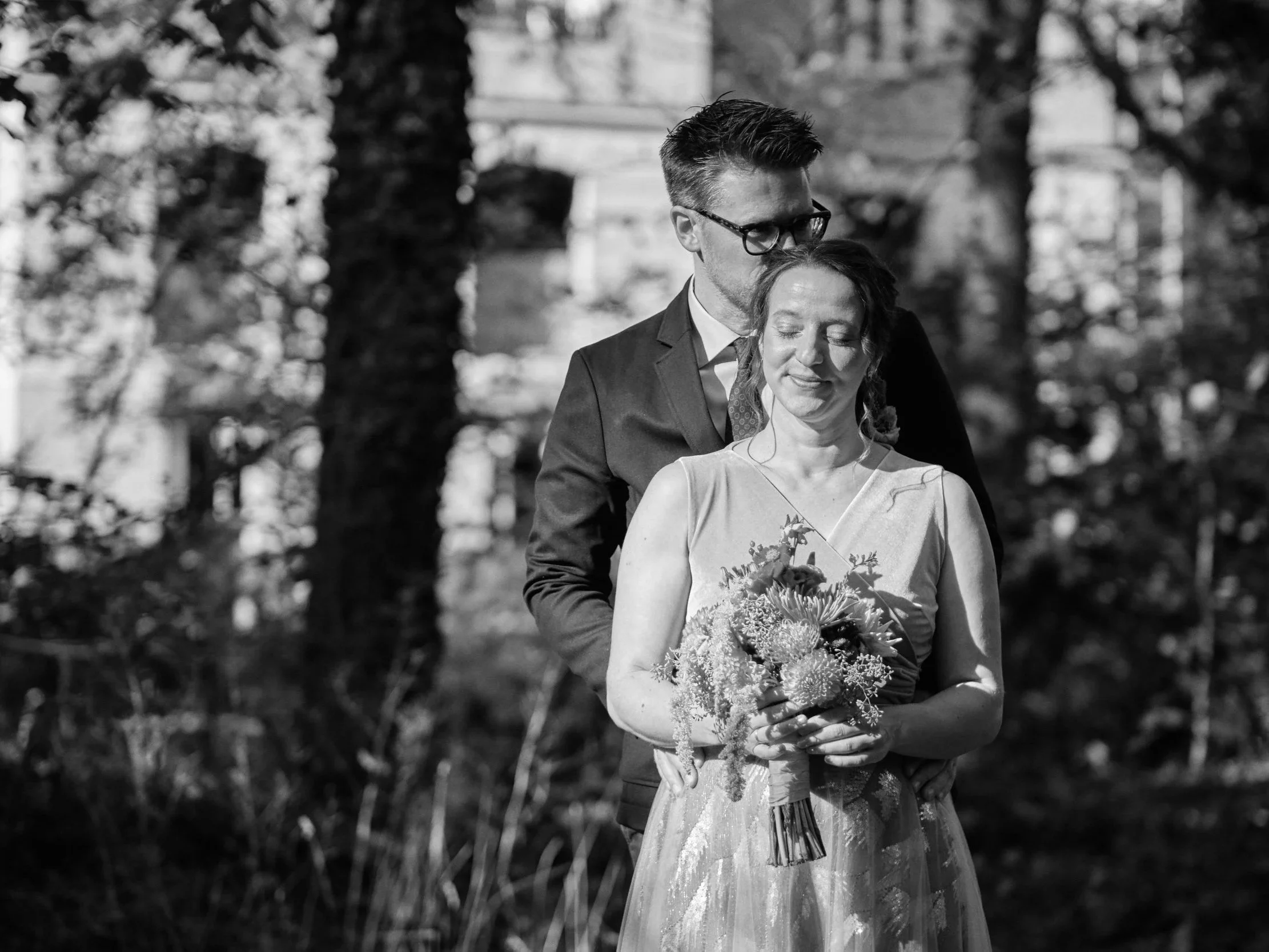 Ein Paar bei einer Hochzeit im Freien, der Mann umarmt die Frau von hinten und küsst sie auf den Kopf, die Frau hält einen Blumenstrauß.
