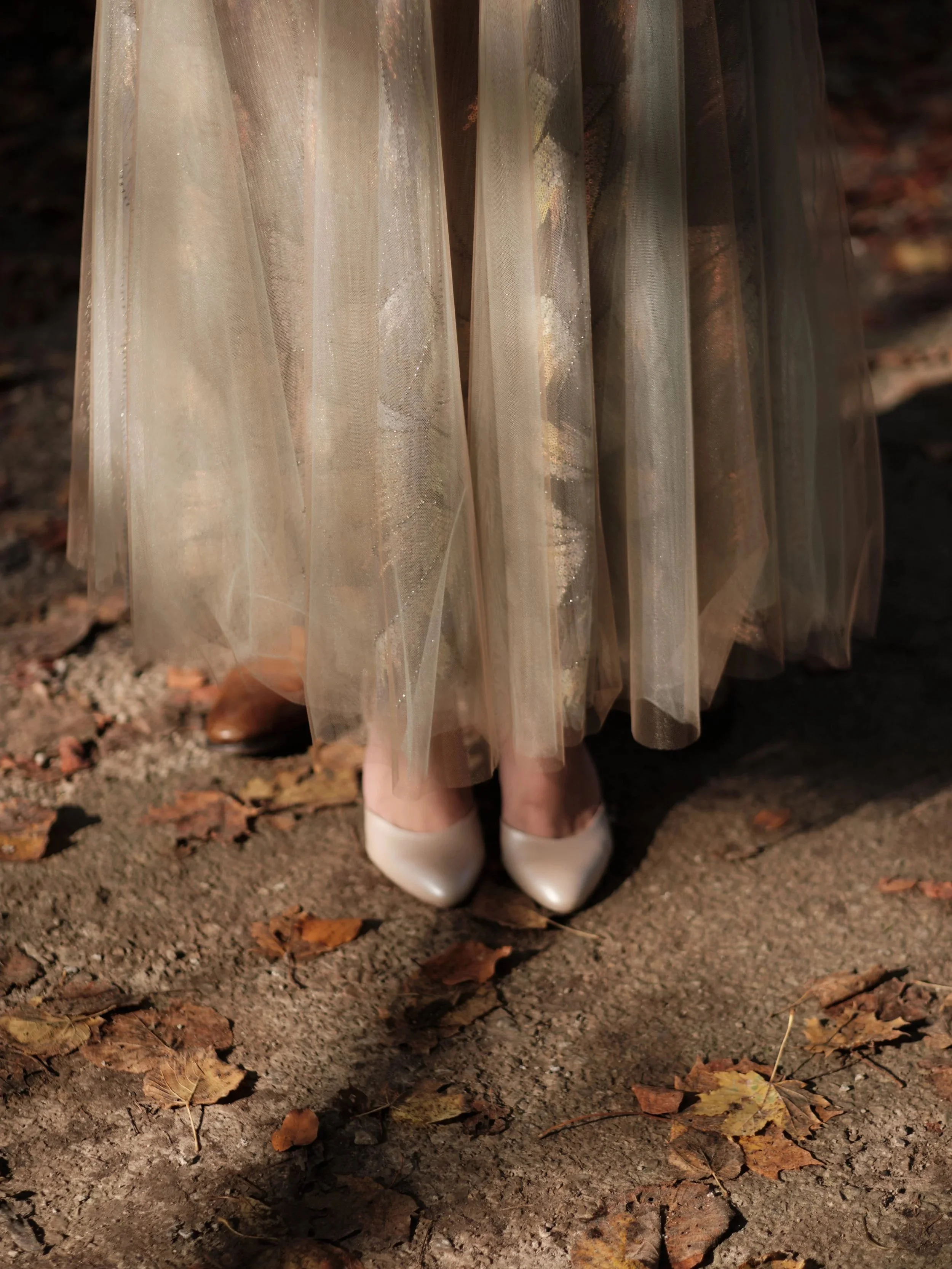 Blick auf die Hochzeitsschuhe der Braut in einem langen, durchsichtigen, glitzernden Tüllkleid und weißen High Heels, auf Erde mit herabfallenden Herbstblättern.