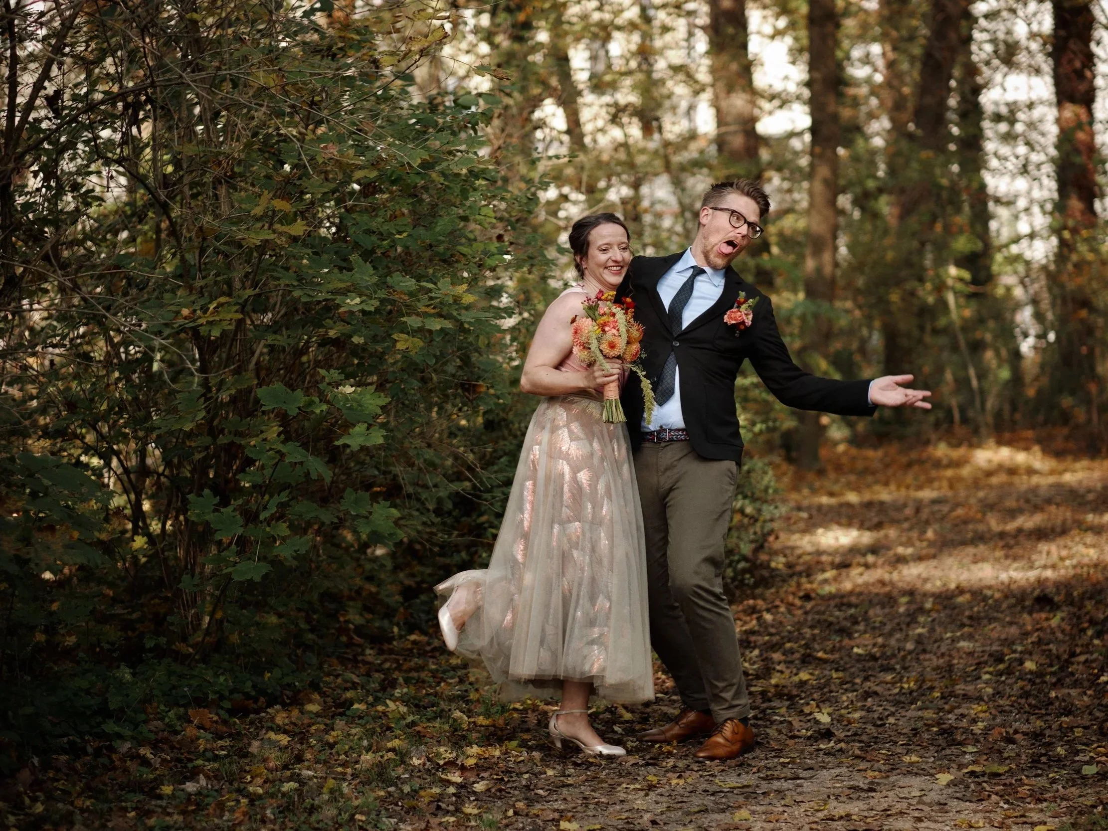 Ein Paar bei einer Hochzeit im Park, die Frau hält einen Blumenstrauß, beide lachen und tanzen, umgeben von herbstlicher Natur.