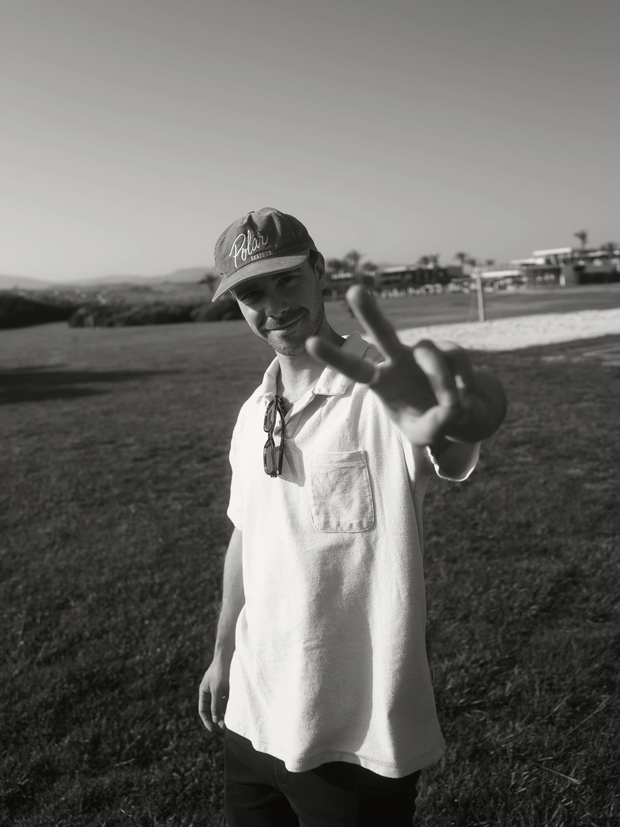 Mann trägt Kappe mit Aufschrift 'Polar' und posiert auf einem Golfplatz, zeigt die Hand in Friedenszeichen, im Hintergrund Bäume und Gebäude, schwarz-weiß Foto.