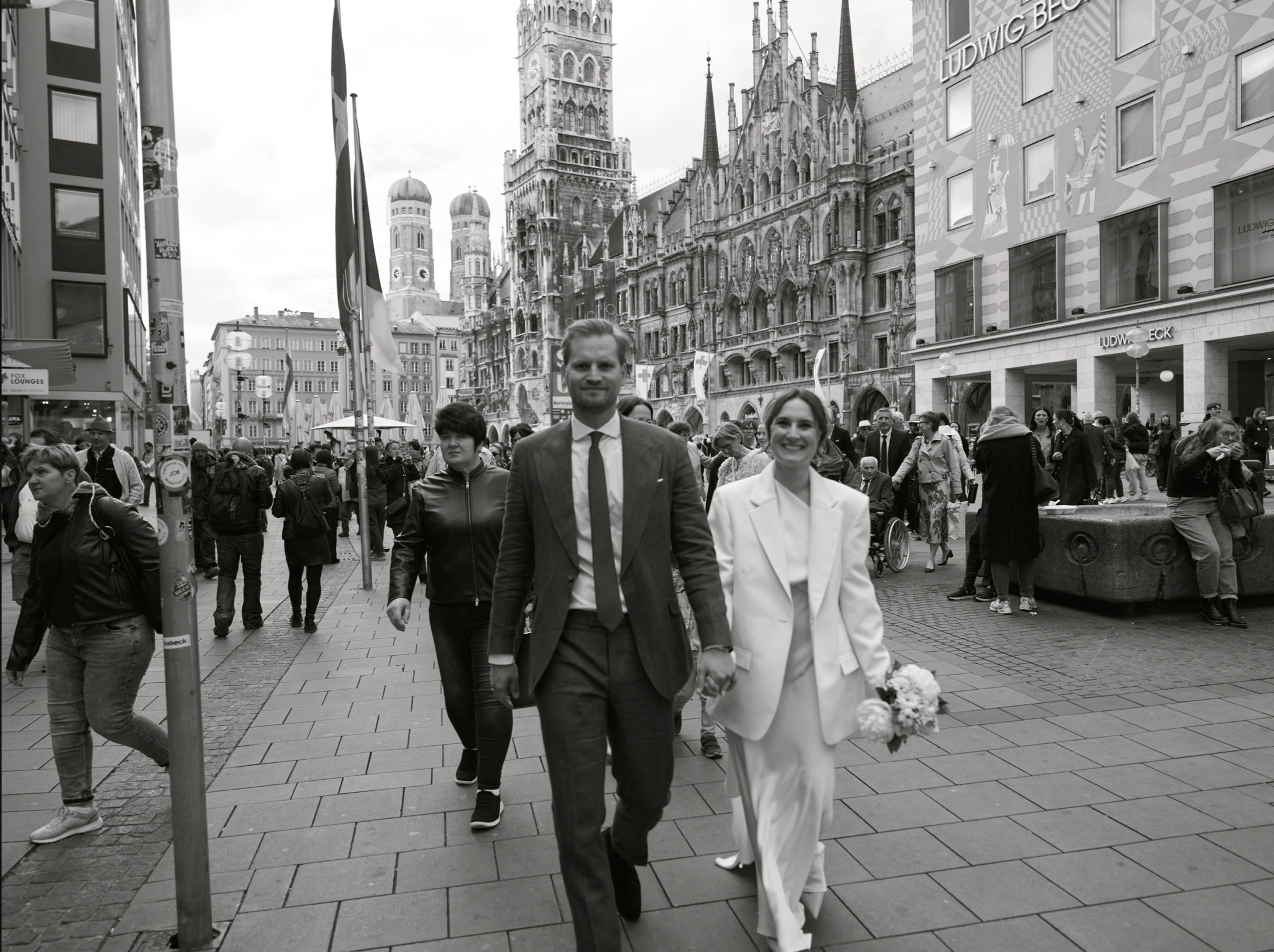 Ein braun-weißesFoto eines Paares, das bei einer Hochzeitsfeier in einer Stadt mit historischen Gebäuden spazieren geht. Das Paar ist in formeller Kleidung, die Frau hält einen Blumenstrauß.