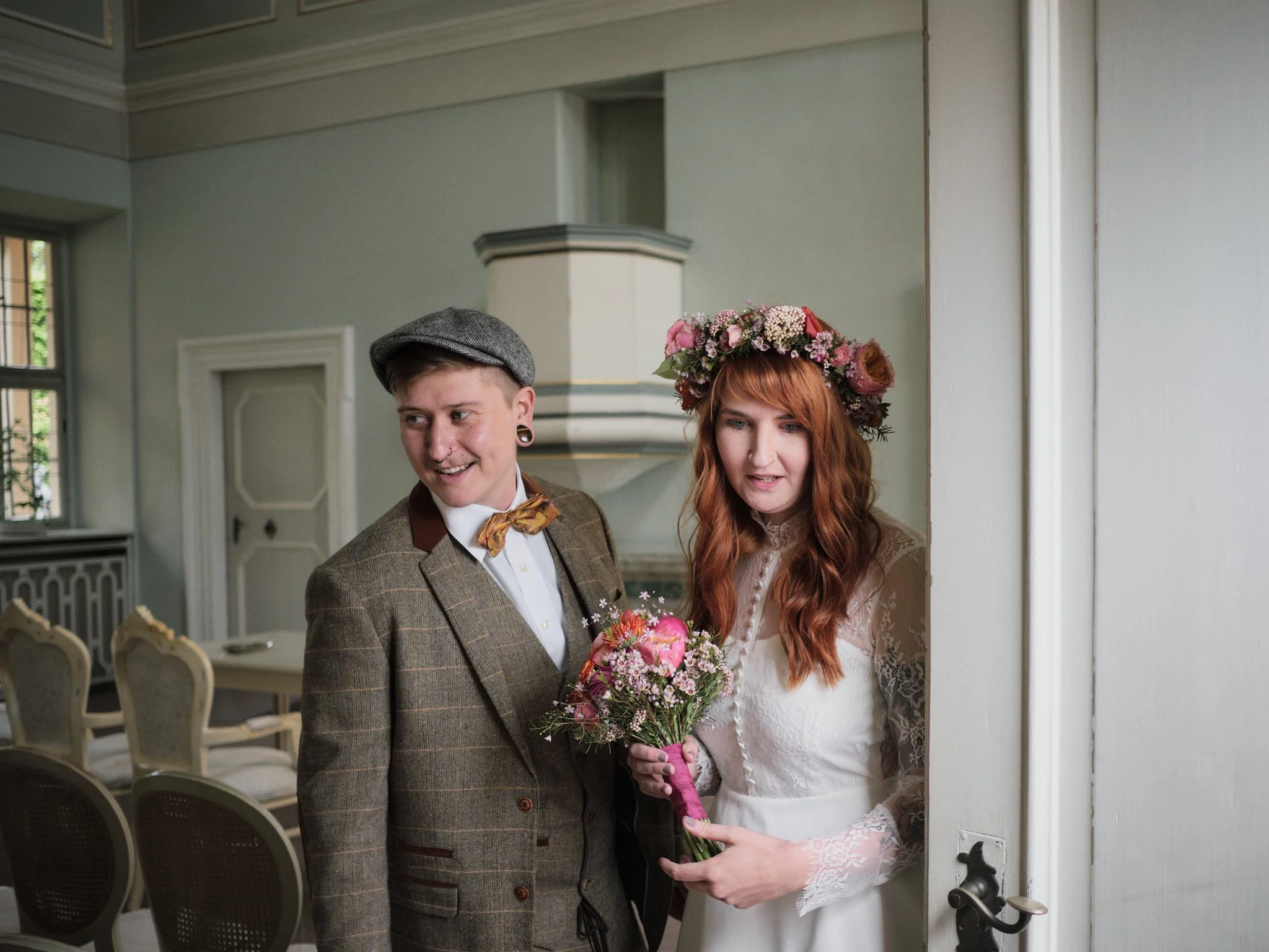 Ein Brautpaar bei einer Hochzeit, die Braut hat rote Haare, trägt ein weißes Brautkleid mit Spitzenärmeln und einen Kranz aus Blumen im Haar, die Braut hält einen Blumenstrauß, der Bräutigam hat einen karierten Anzug und trägt ein Grau-Fedora, sie st