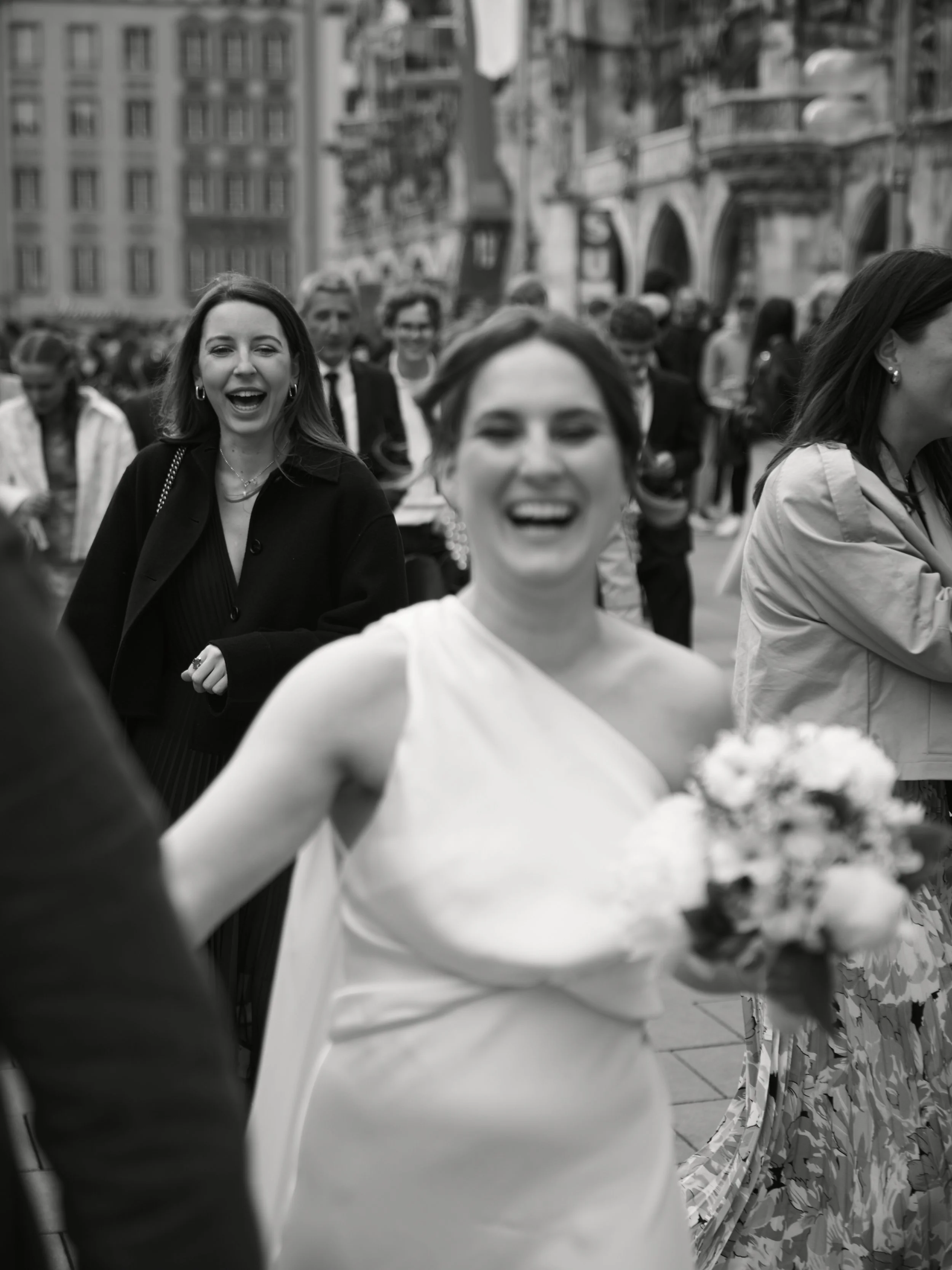 Frauen lachen und feiern auf einer Straße, die Frau trägt ein weißes Kleid und hält einen Blumenstrauß, im Hintergrund sind weitere Menschen und alte Gebäude.