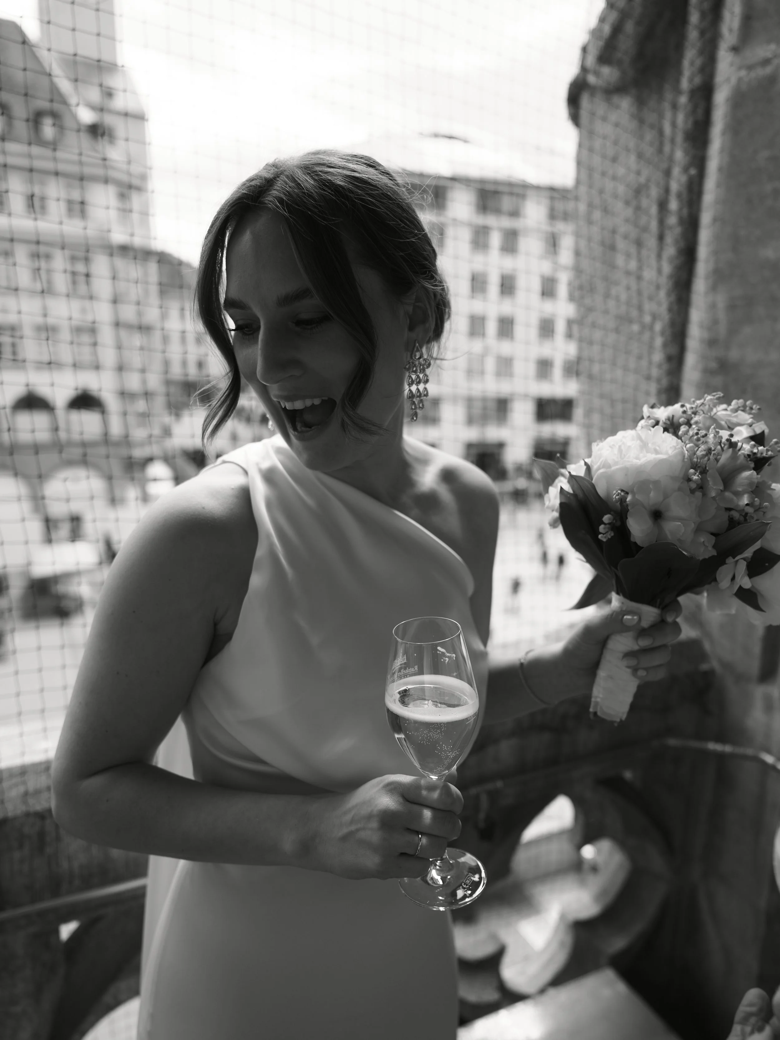 Die Braut trägt ein elegantes weißes Kleid, hält ein Glas mit Sekt in der einen Hand und einen Blumenstrauß in der anderen auf einem Balkon, im Hintergrund ist der Marienplatz in München zu sehen.