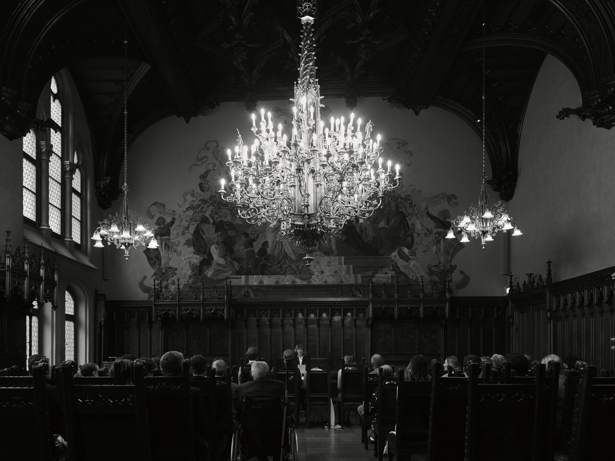 Ein eleganter, historischer Sitzungssaal mit großen Fenstern, Wandgemälden, einer großen Kristallleuchter, Menschen in formeller Kleidung und einem Redner am Podium.