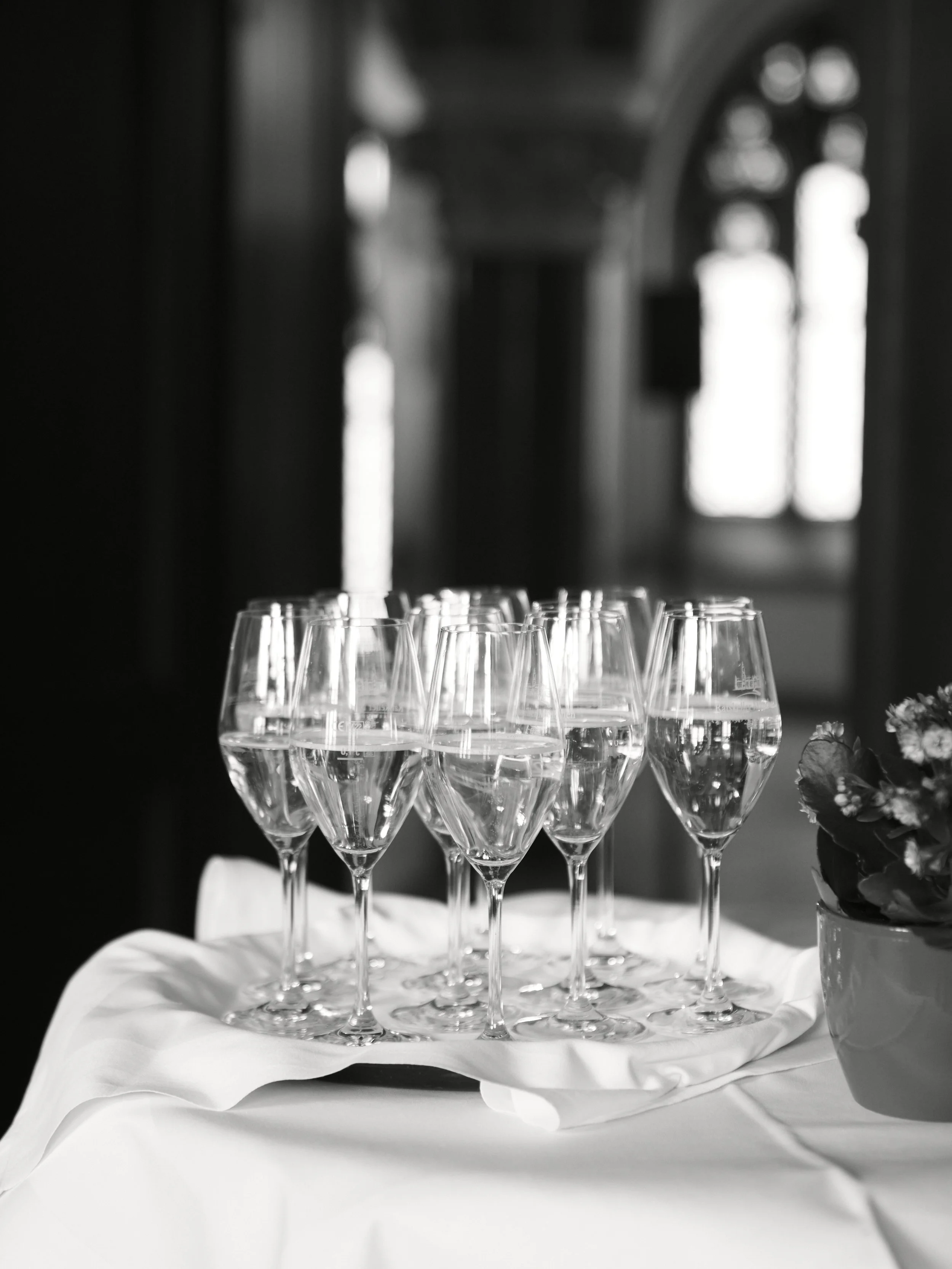 Sechs Sektgläser auf einem weißen Tuch bei einer Feier im eleganten Raum mit hohen Fenstern in Schwarz-Weiß aufgenommen.