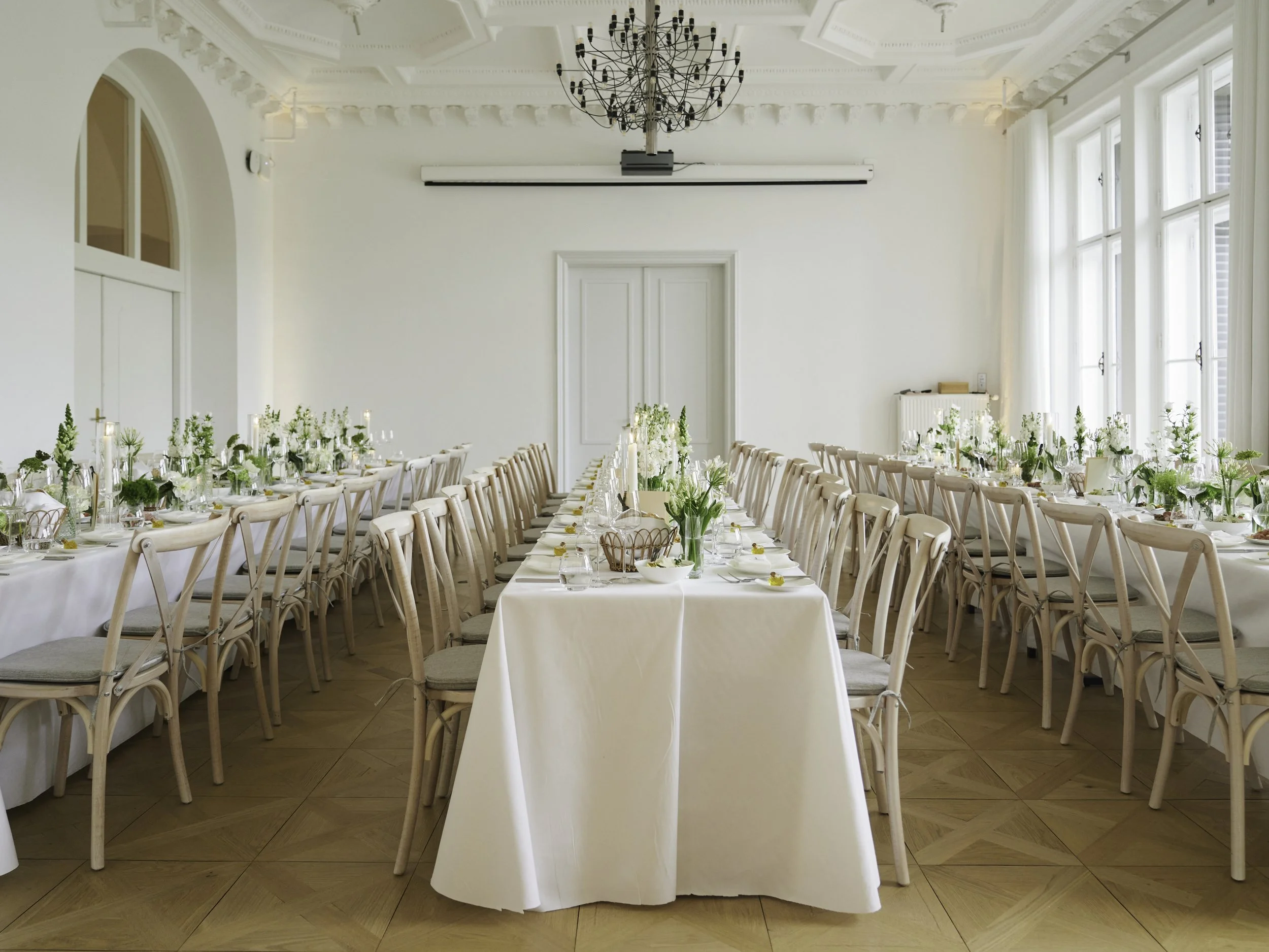Ein eleganter festlich gedeckter Saal mit mehreren langen Tischen, weißem Tischtuch, weißen Blumenarrangements und Kerzen, große Fenster lassen viel Tageslicht herein, im Hintergrund eine große weiße Tür, die Decke ist mit kunstvollen Stuckdetails ve