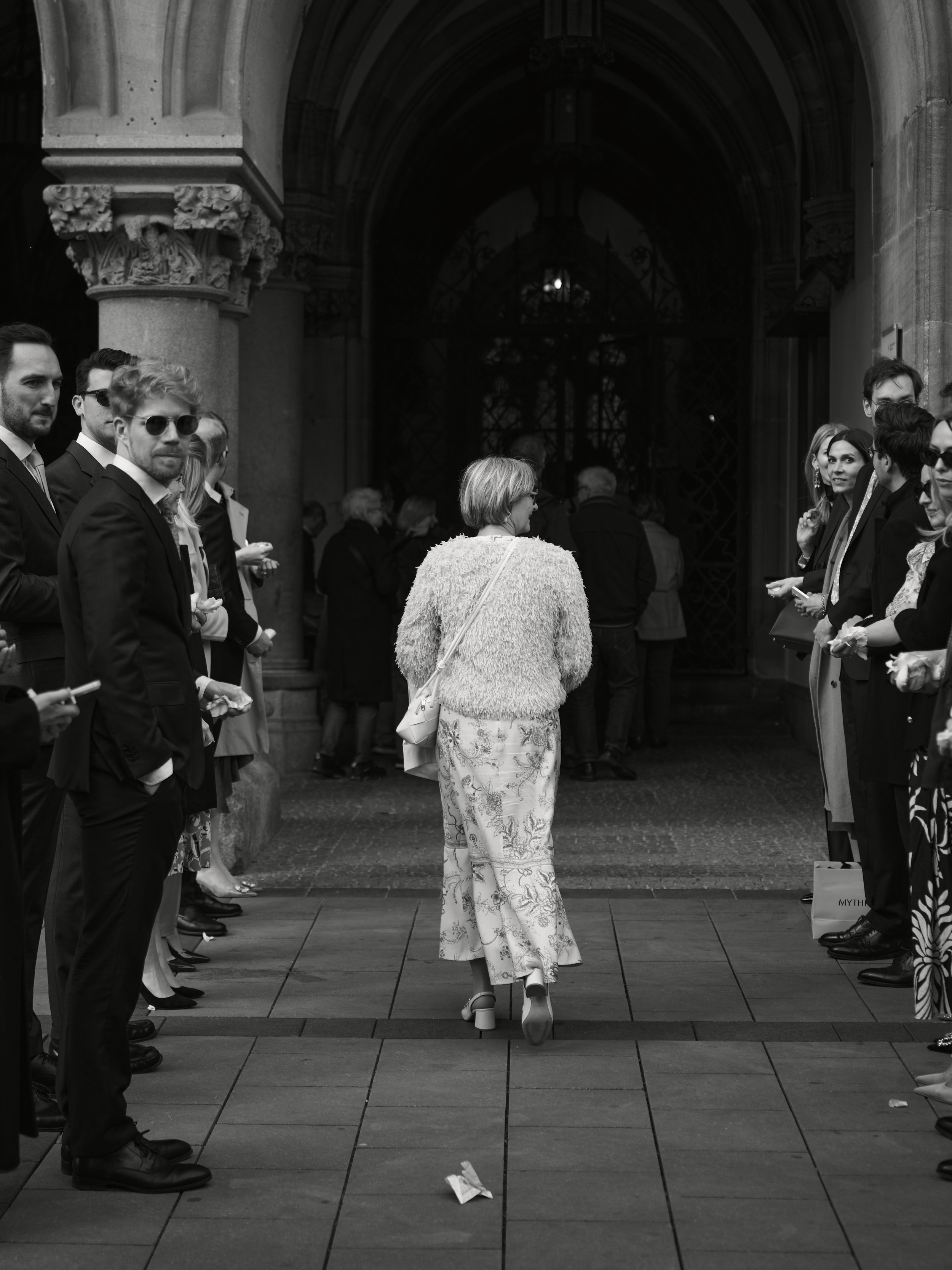Frau mit einem abgenutzten breitkrempigen Hut läuft durch eine Menschenmenge vor einem historischen Gebäude. Die Szene ist schwarz-weiß aufgenommen.