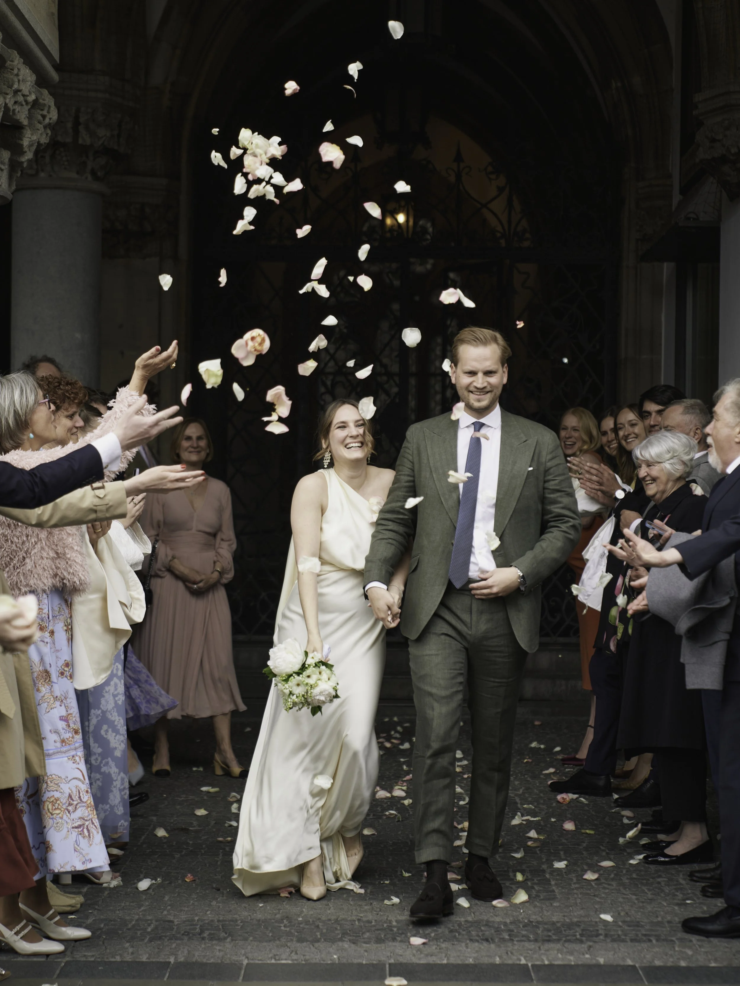 Ein glückliches Brautpaar beim Verlassen eines Gebäudes, umgeben von jubelnden Freunden und Familie, während Blumenblätter in die Luft geworfen werden.