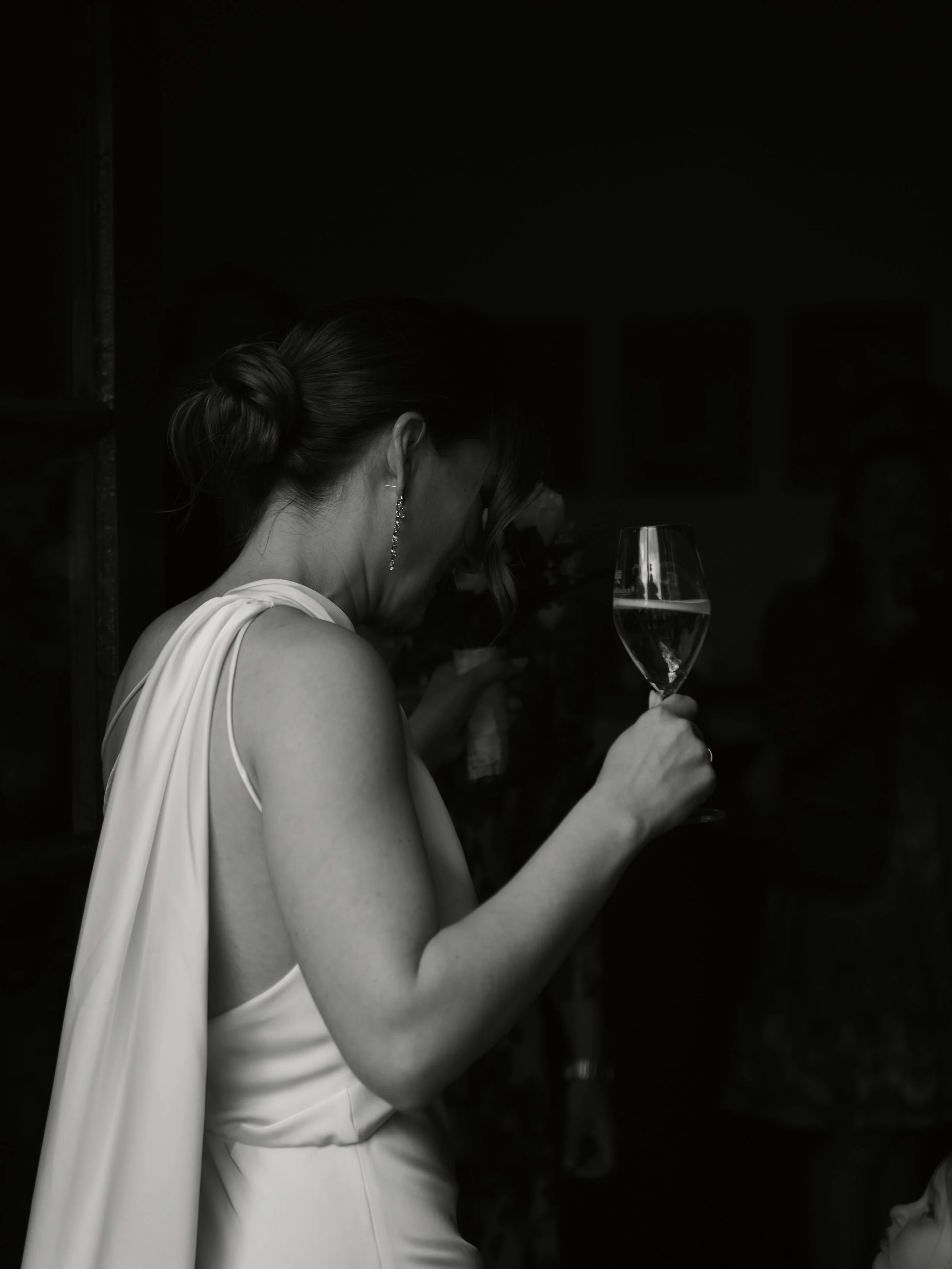 Braut in einem weißen Kleid hält ein Glas Sekt bei ihrer Hochzeitsfeier, im Hintergrund sind andere Menschen in einem dunklen Raum.