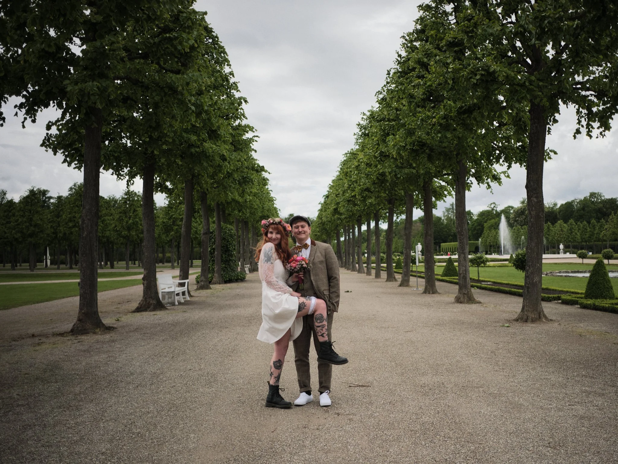 Ein lachendes Hochzeitspaar steht in einem Schlosspark mit einer Lindenallee, einer Springbrunnen und gepflegten Hecken im Hintergrund.