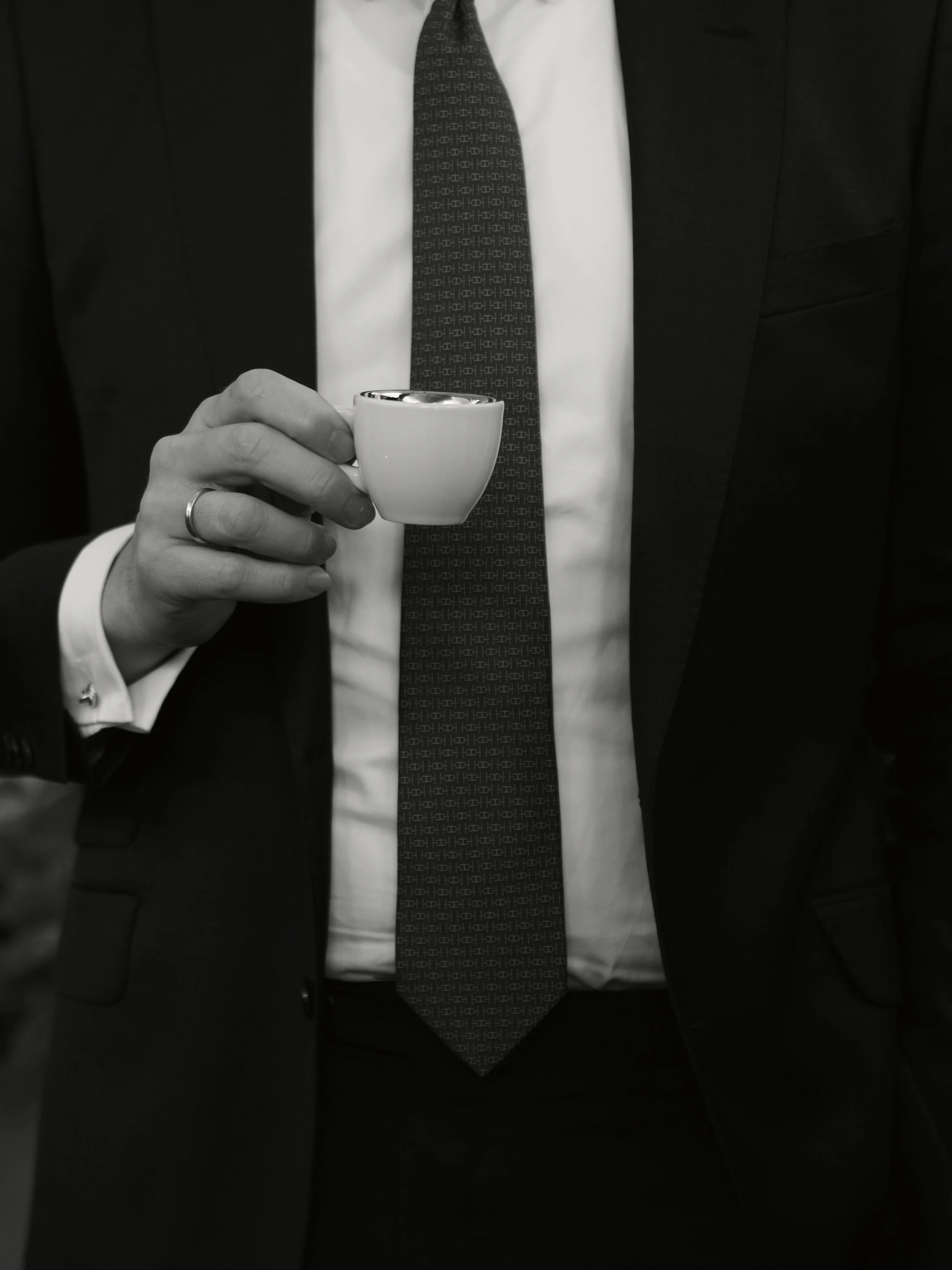 Ein Mann in formeller Kleidung hält eine kleine weiße Tasse. Er trägt einen schwarzen Anzug, weißes Hemd und eine dunkle Krawatte mit einem geometrischen Muster.