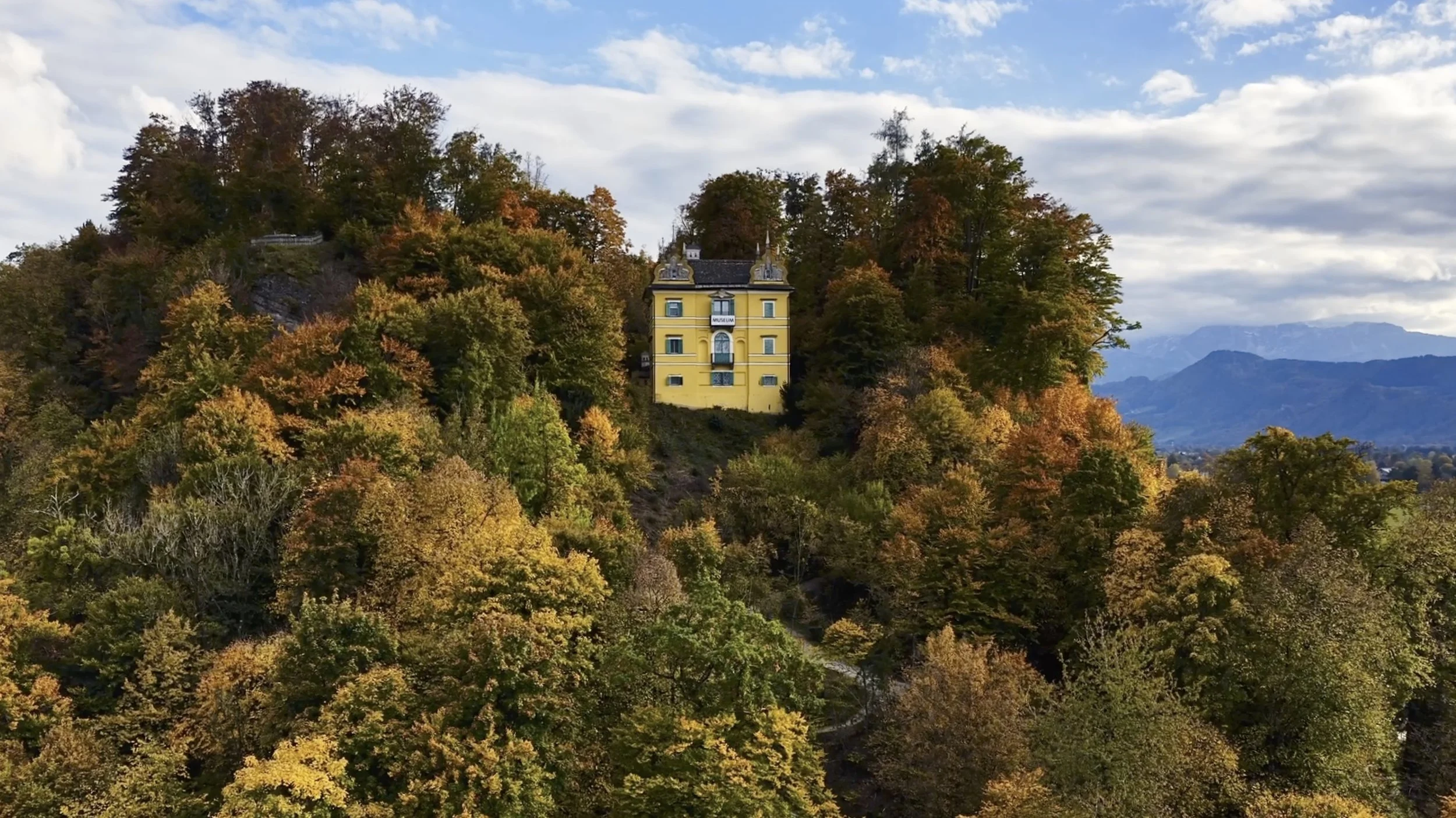 Ein gelbes Haus auf einem Hügel, umgeben von buntem Herbstwald mit roten, orangefarbenen und grünen Bäumen, mit Bergen im Hintergrund und einem blauen Himmel mit Wolken.