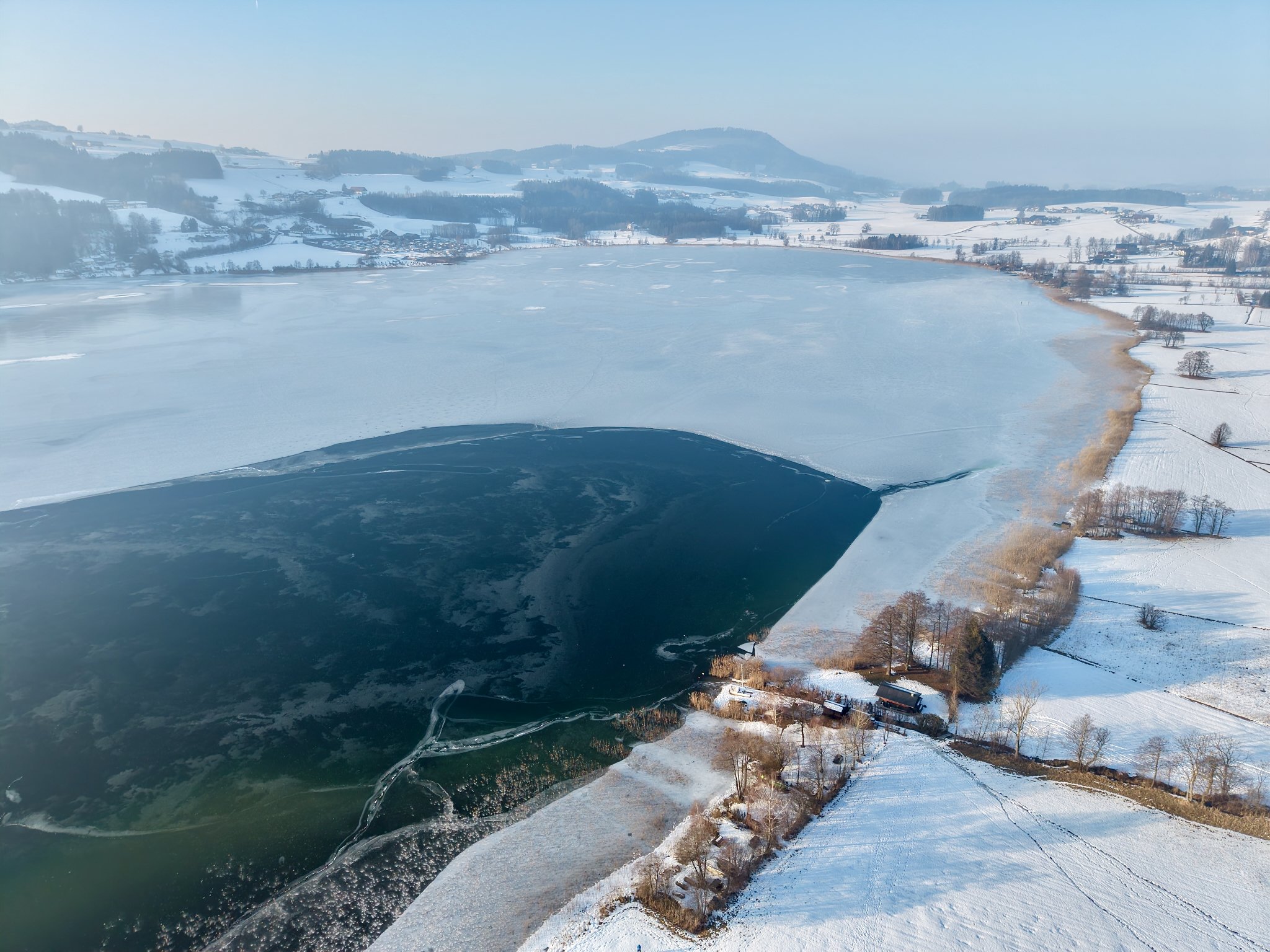 Vogelperspektive auf einen gefrorenen See mit Schneeflächen und umliegenden verschneiten Landschaften sowie Bäume und Häuser im Hintergrund.