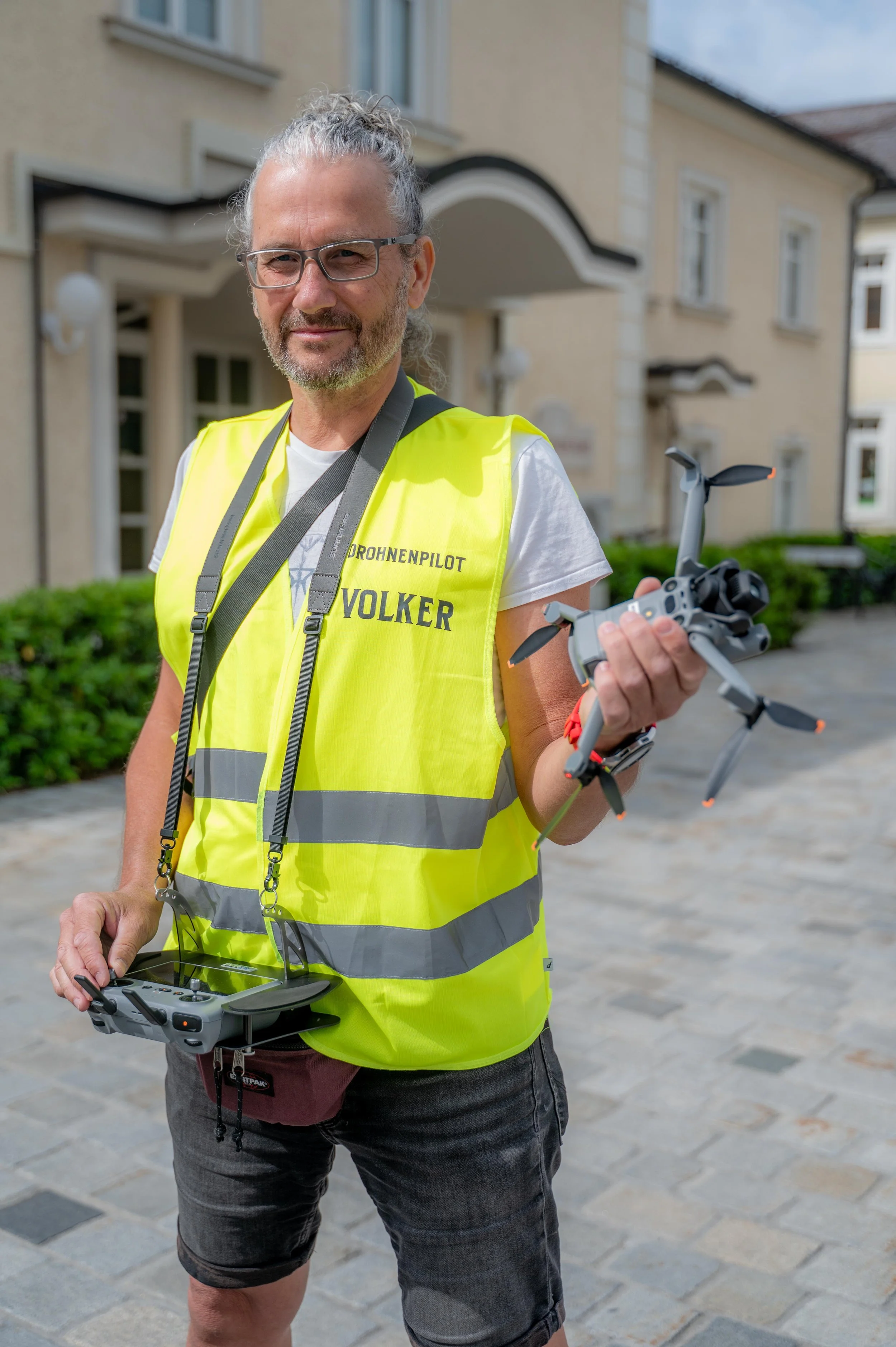 Mann trägt gelbe Warnweste mit der Aufschrift 'Drohnenpilot Volker', hält eine Drohne in der Hand und steuert sie mit einem Fernsteuergerät, steht vor einem Wohnhaus im Freien.