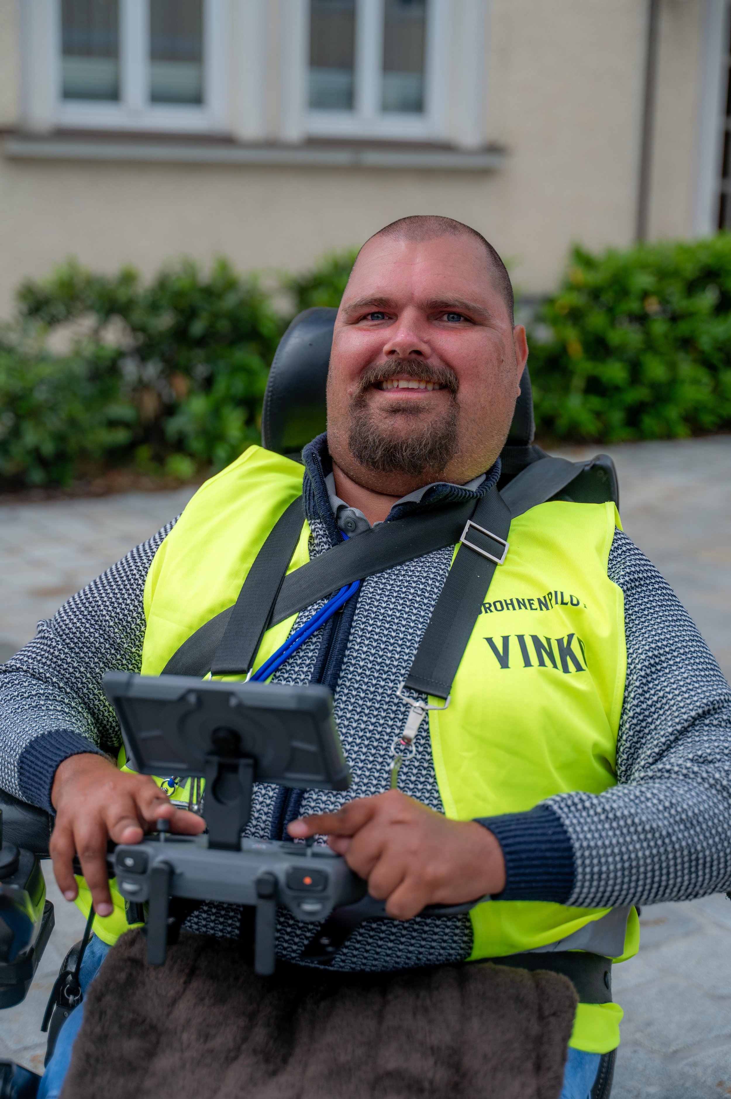 Mann im Rollstuhl mit gelber Sicherheitsweste, der mit einem Drohnen-Controller arbeitet, vor einem Haus mit Fenstern und Gehweg, im Freien.