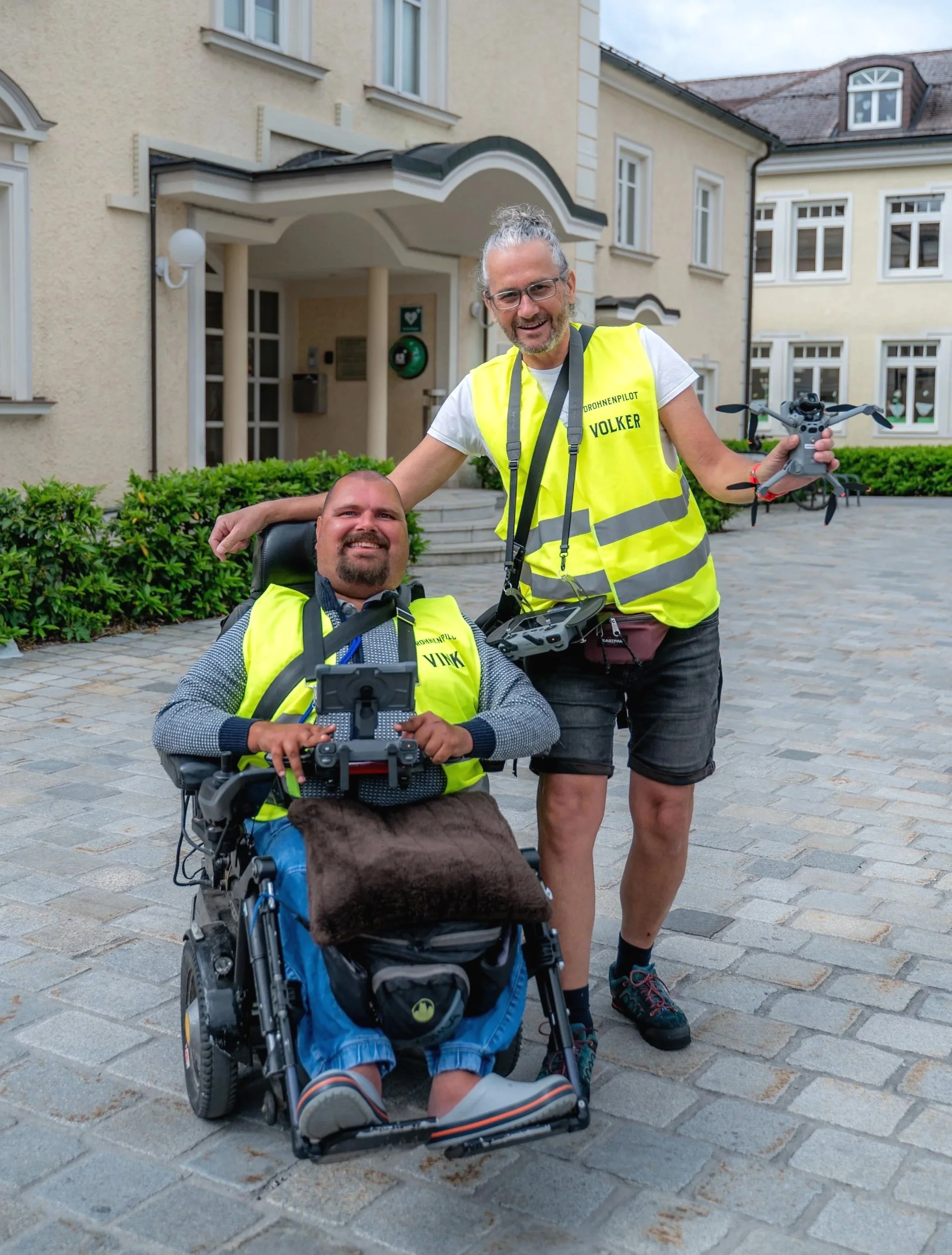 Zwei Männer, einer im Rollstuhl, beide in leuchtend gelben Sicherheitswesten, vor einem großen Gebäude. Der stehende Mann hält eine Drohne und lacht, der sitzende Mann hält eine Fernbedienung für den Rollstuhl und trägt ebenfalls eine Sicherheitsweste.
