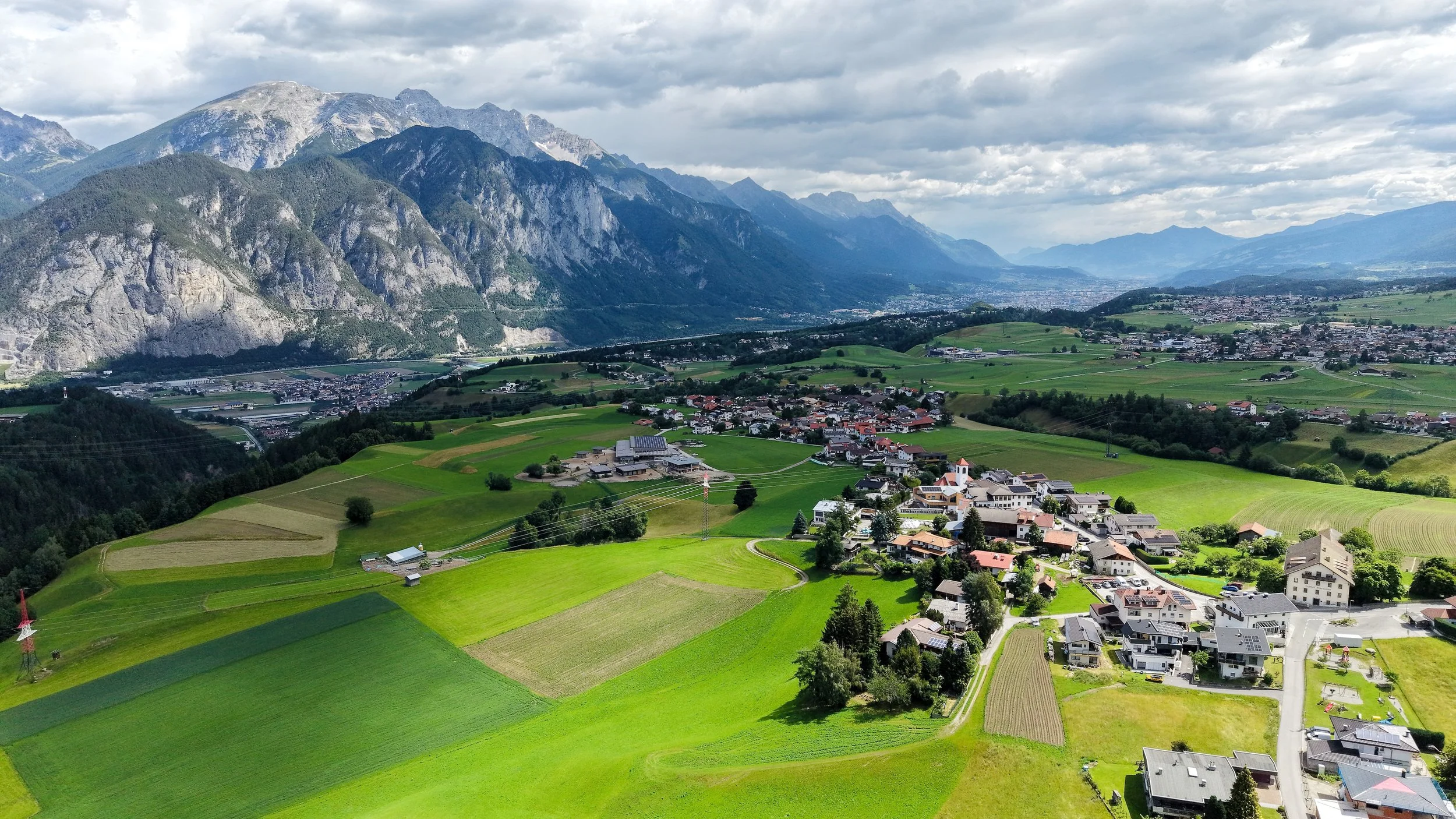 Landschaft mit grünen Feldern, kleinen Häusern, Bergen im Hintergrund und bewölktem Himmel.
