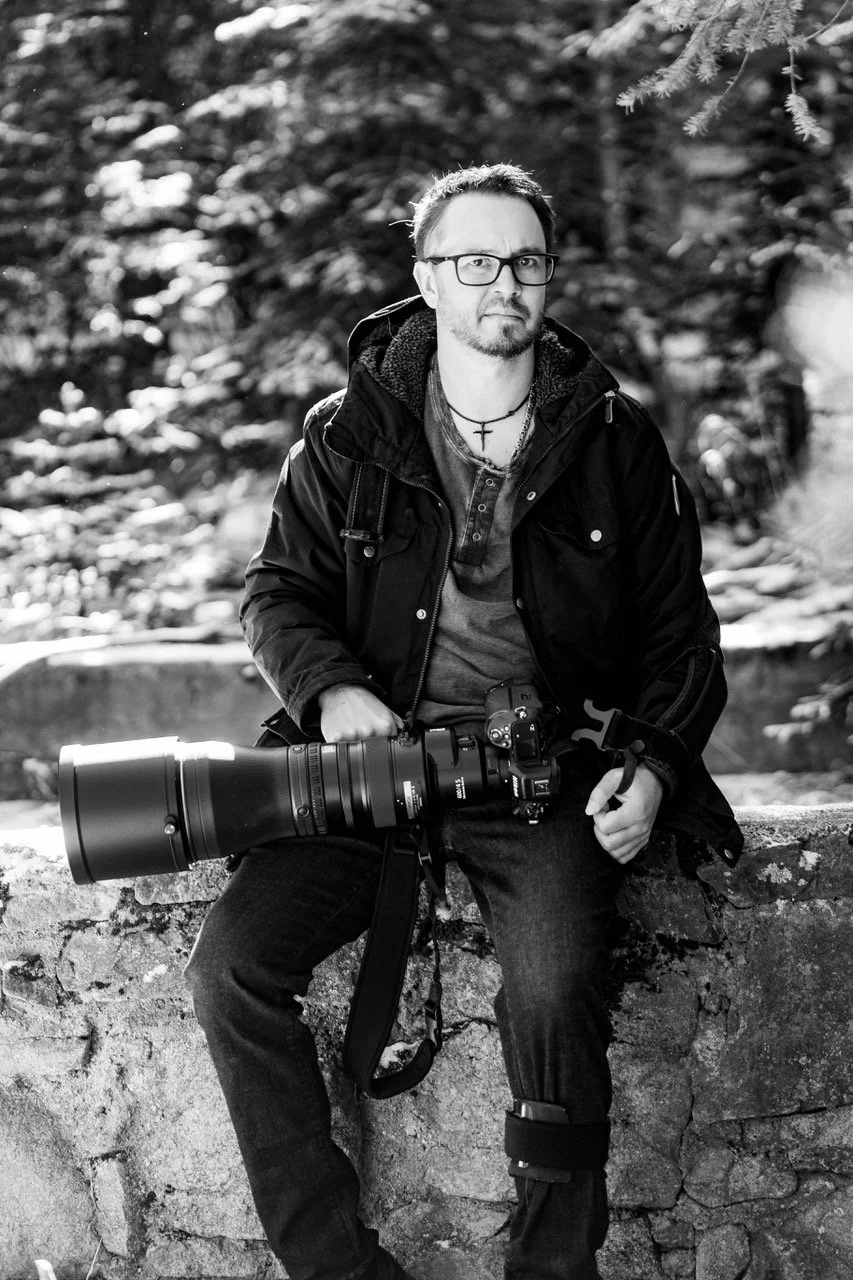 Bryan Fevold with glasses, a beard, and a cross necklace, holding a large camera with a telephoto lens, sitting on a stone wall outdoors in a wooded area.