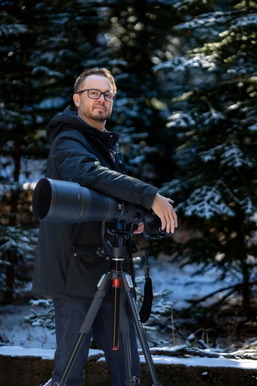 Bryan Fevold with glasses and a beard stands outdoors in a winter forest, holding a large camera with a telephoto lens mounted on a tripod.