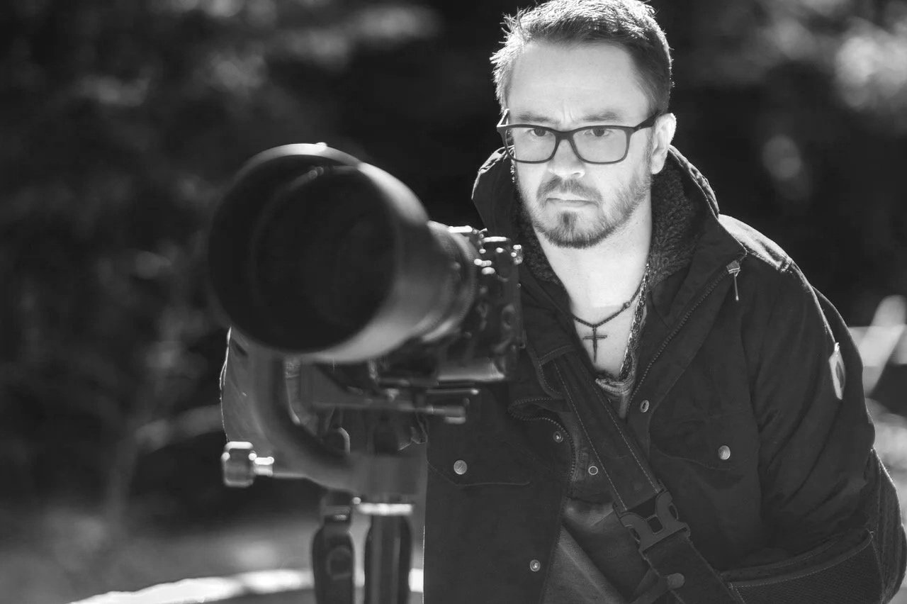 Bryan Fevold with glasses and a beard looking through a telescope outdoors in black and white.