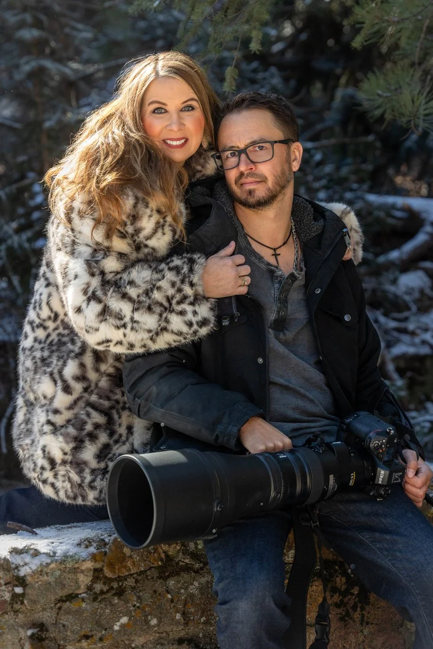 Bryan Fevold's wife with long, wavy blonde hair wearing a leopard print coat standing next to a Bryan Fevold with short dark hair, glasses, and a beard, dressed in dark clothing with a camera around his neck, seated outdoors in a winter wooded area.