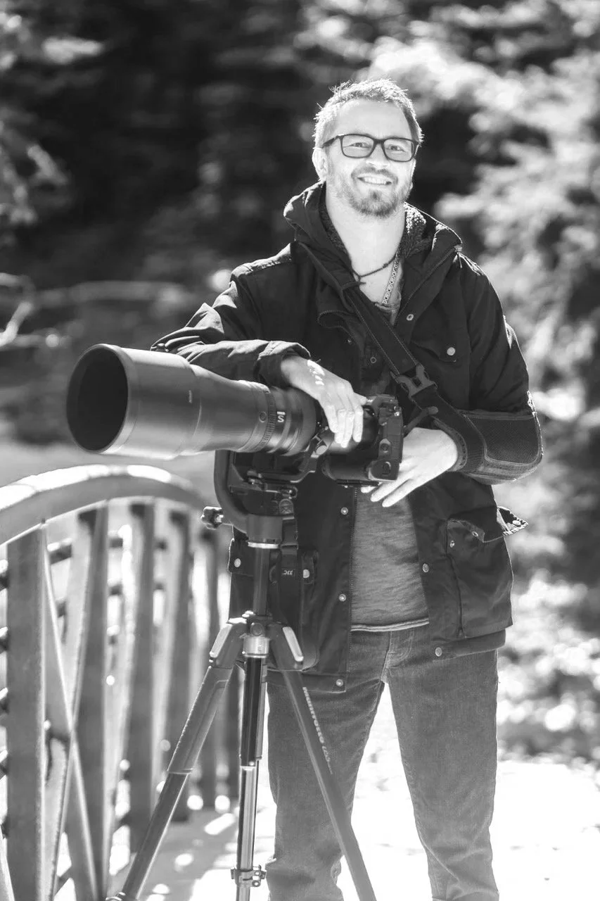 Bryan Fevold with glasses and a beard stands outdoors, smiling, holding a camera with a large telephoto lens mounted on a tripod.