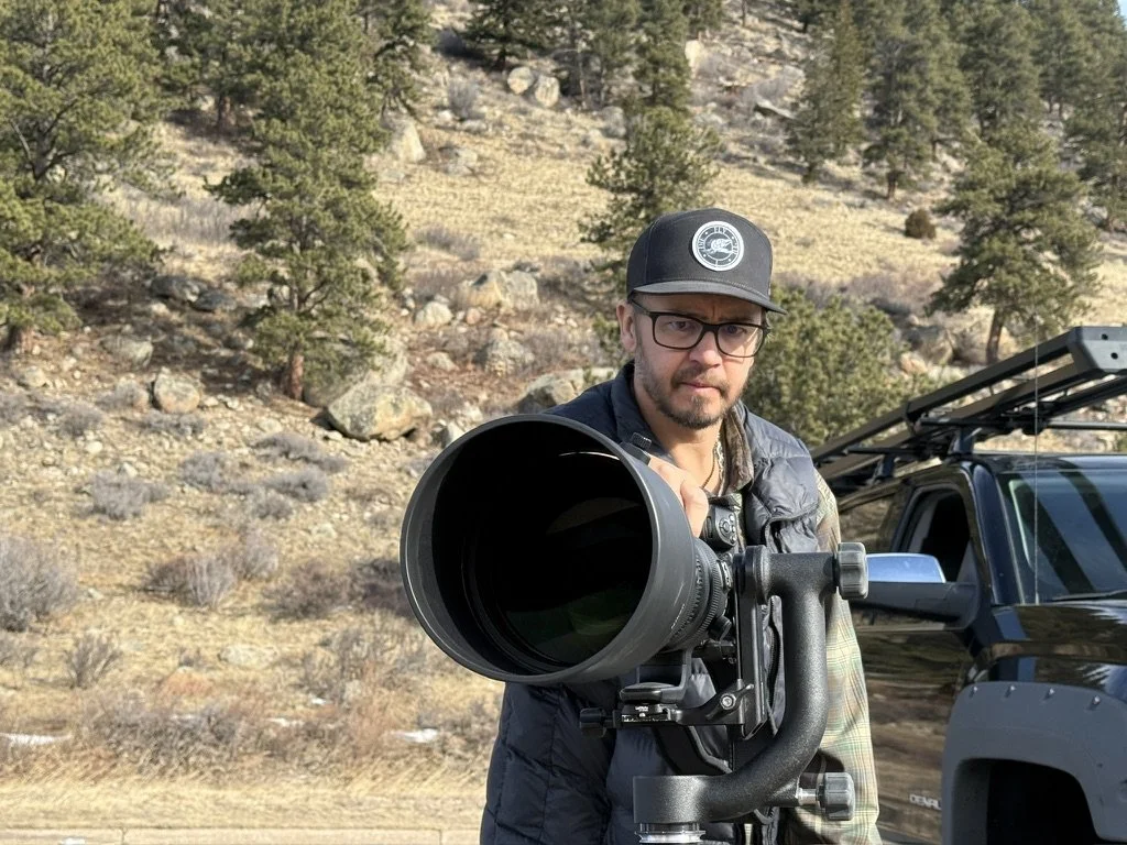 Bryan Fevold wearing glasses, a black cap, and outdoor clothing is looking through a large telescope outdoors in a forested area.