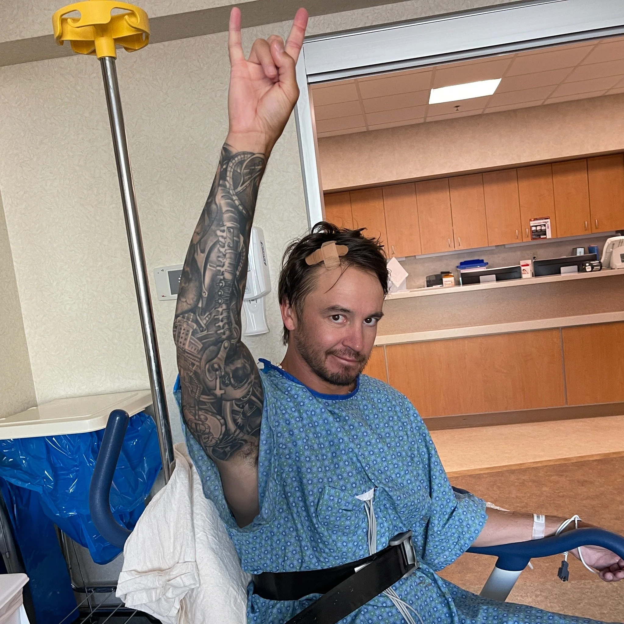 A man in a hospital gown sitting in a hospital bed, raising his right hand with two fingers up in a peace sign, with a bandage on his forehead.