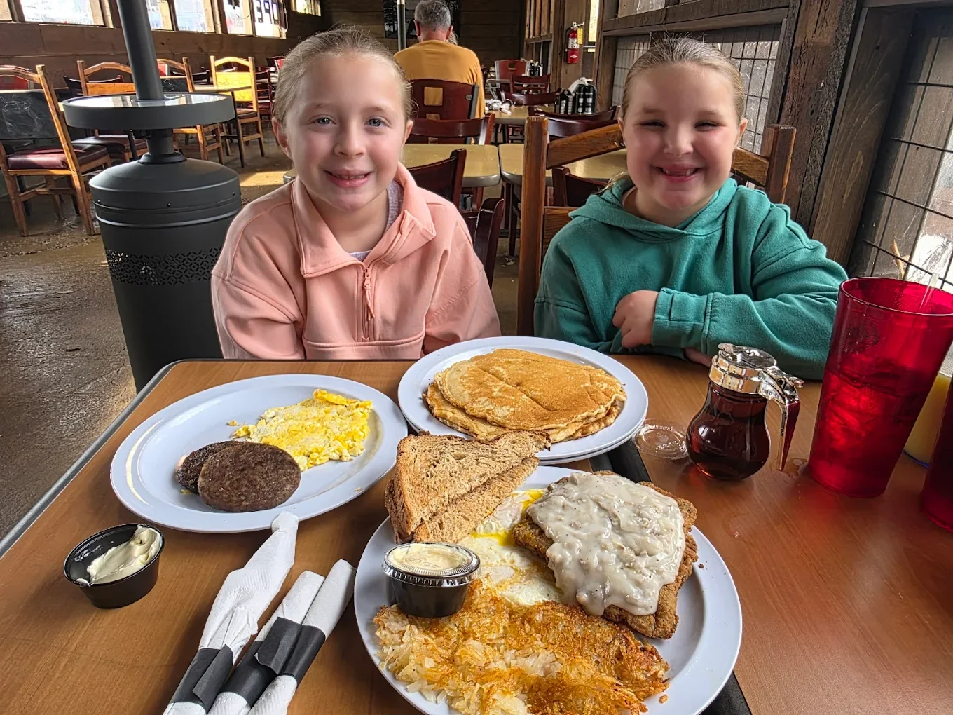 Two young girls sitting at a restaurant table with breakfast food, smiling. The table has plates of scrambled eggs, sausage patties, toast, hash browns with gravy, and pancakes.