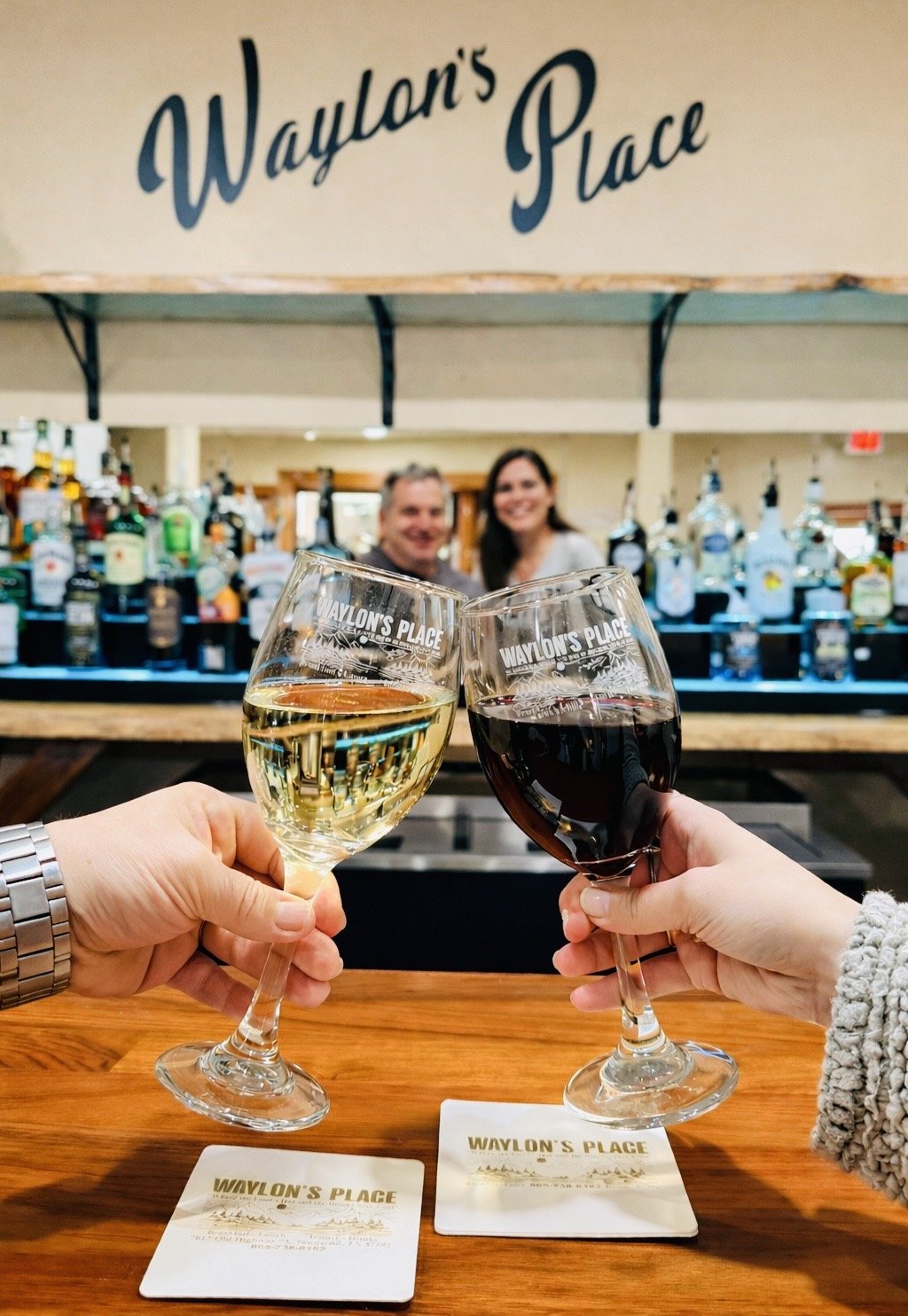 Two people holding glasses of white and red wine in front of a bar with two bartender behind it, and a sign that reads "Waylon's Place" overhead.
