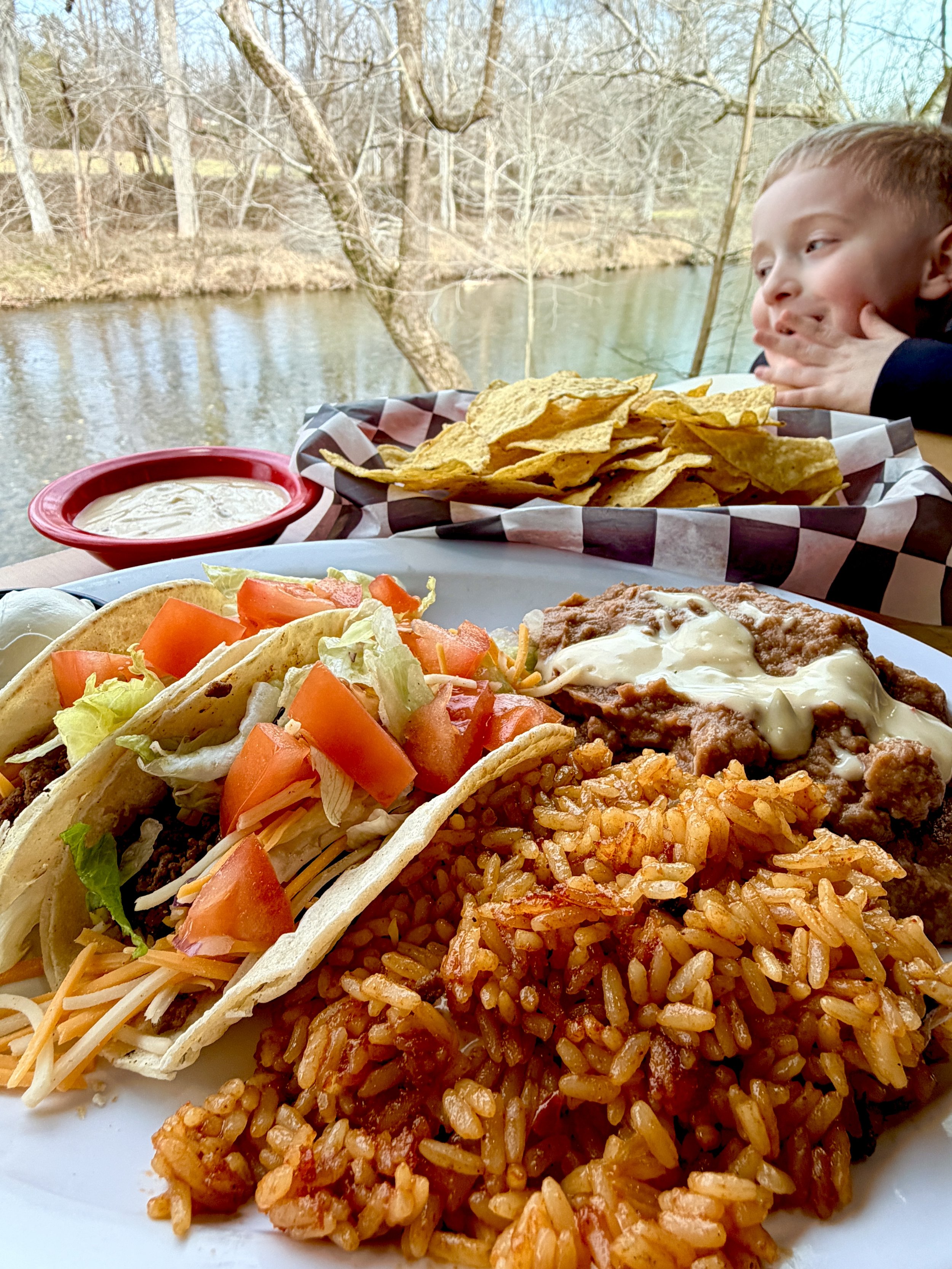 Close-up of a meal with Mexican food, including rice, beef taco with lettuce and tomatoes, a side of refried beans with cheese, tortilla chips, and a small bowl of sauce, outdoors near a river, with a child resting his face on his hands in the backgr