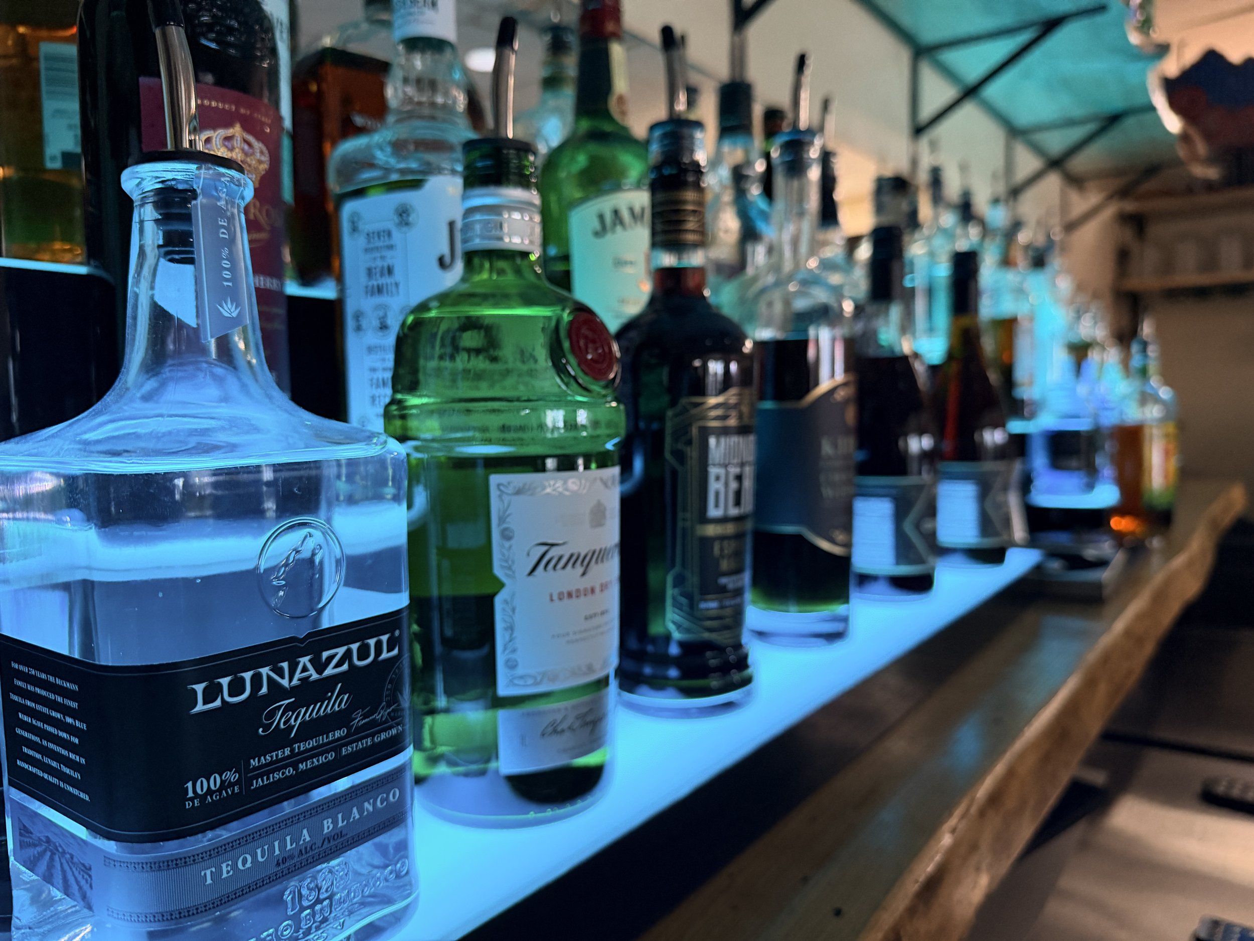 A variety of liquor bottles, including tequila and vodka, displayed on a bar shelf illuminated with blue lighting.